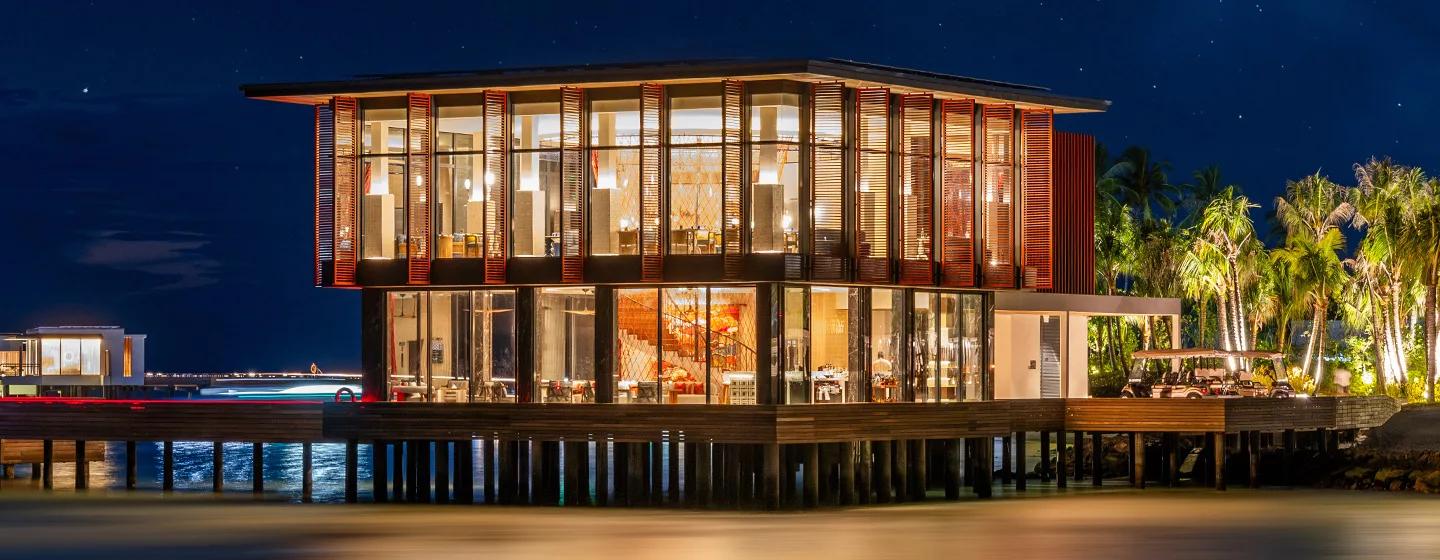 Pavilion Bar's two-level, glass-walled building lit at dusk, built out over the lagoon at SO/ Maldives