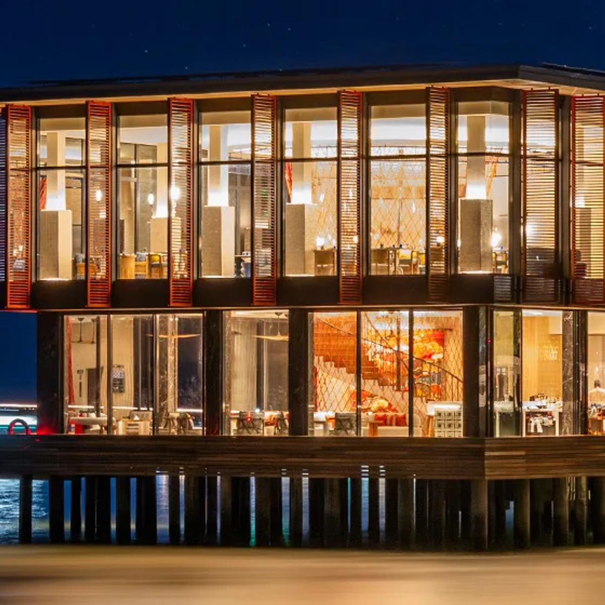 Pavilion Bar's glass-walled, two-level building lit at dusk, built out over the lagoon at SO/ Maldives, with a speedboat's light trail passing below