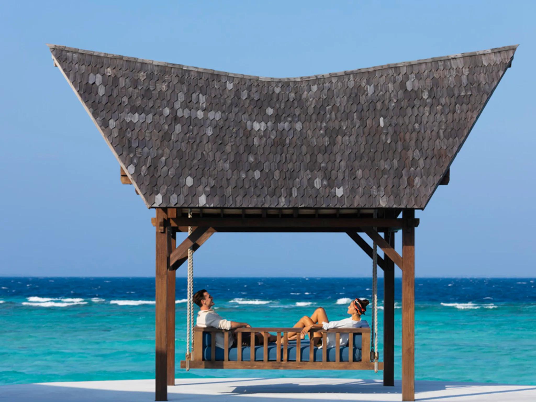 A couple seated on a suspended wooden bench swing beneath a boat-shaped shingled roof structure, open ocean behind them