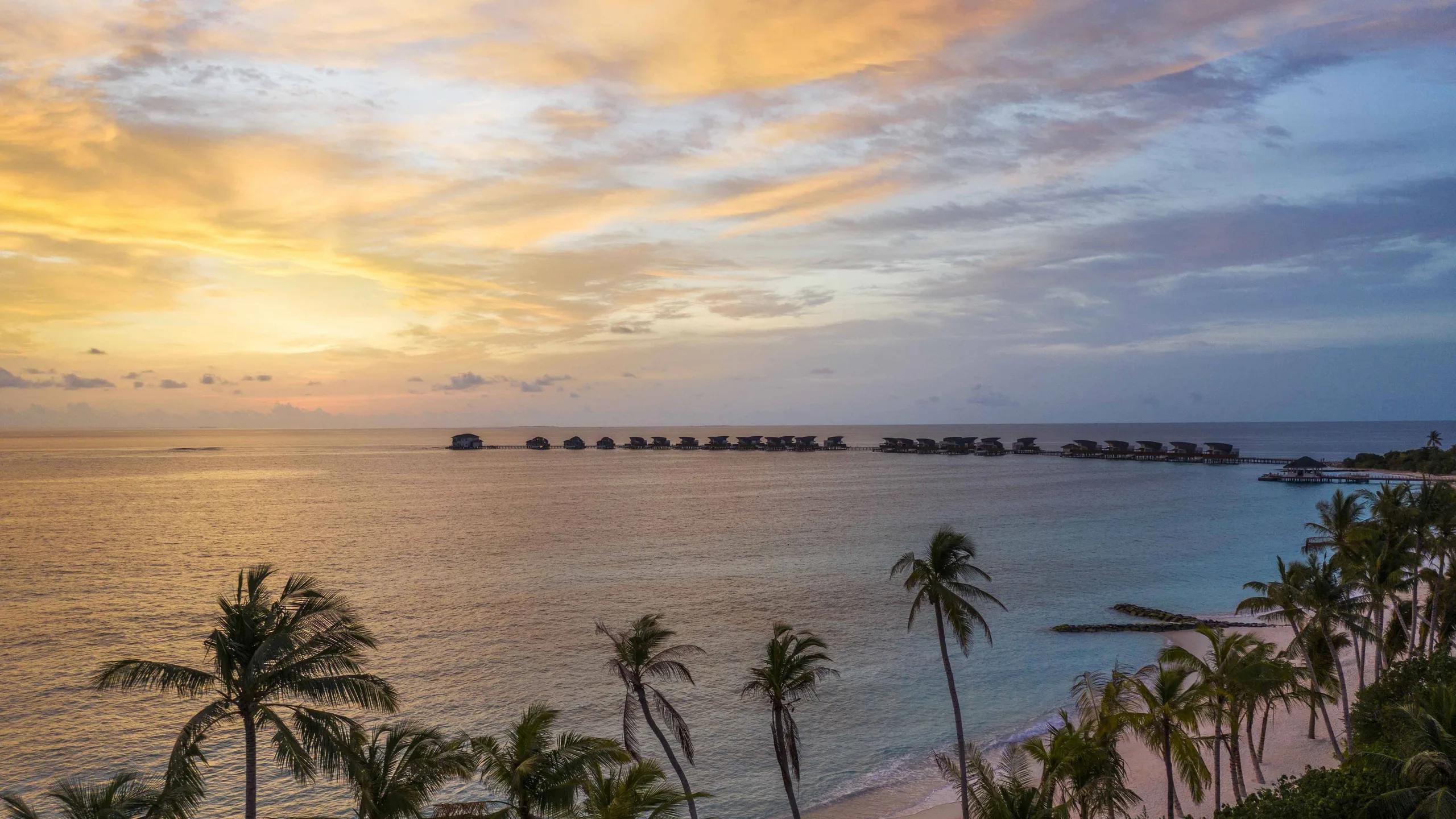 Elevated sunset view over Vagaru Island's overwater villas, palm trees in the foreground
