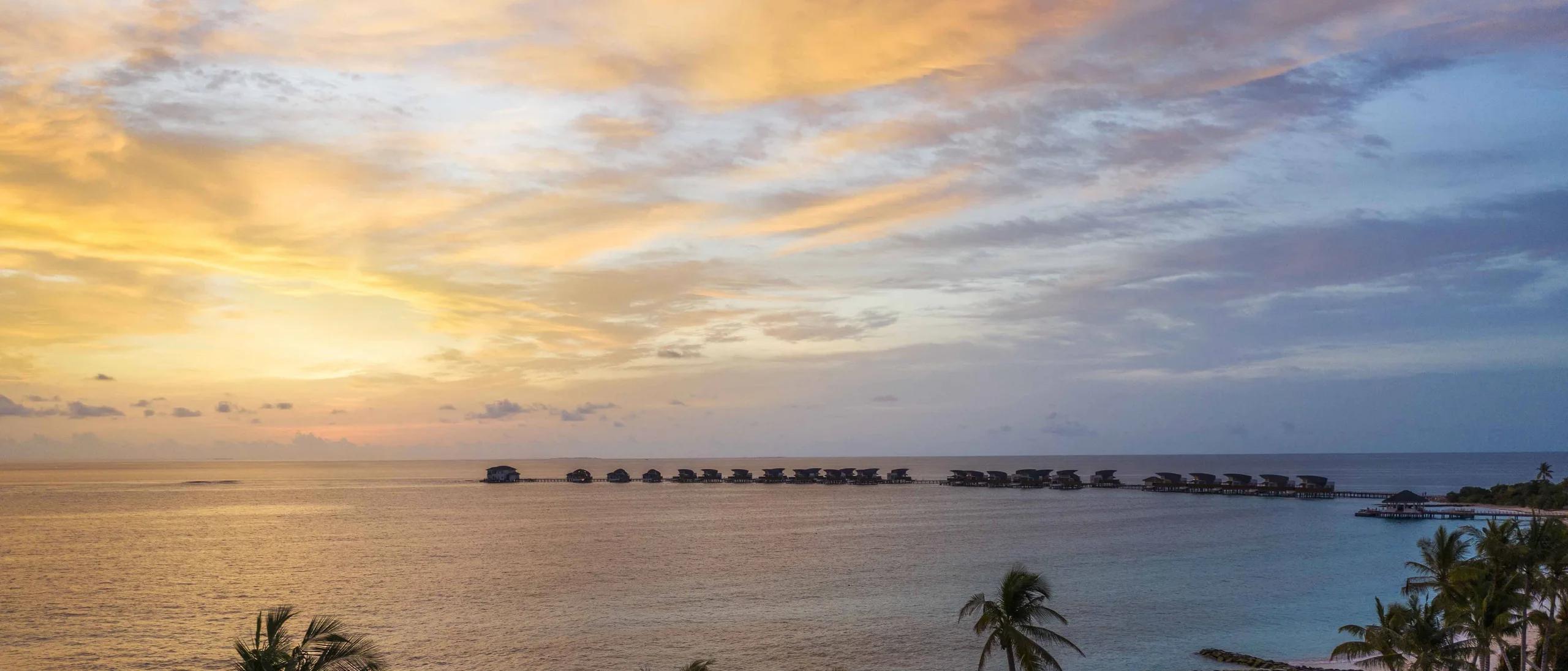 Elevated sunset view over Vagaru Island's overwater villas, palm trees in the foreground