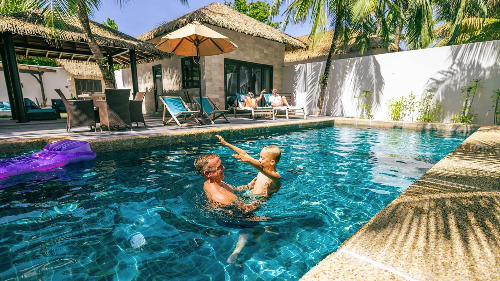 A family swimming together in a Private Pool Villa Family Suite's private pool, thatched roof behind