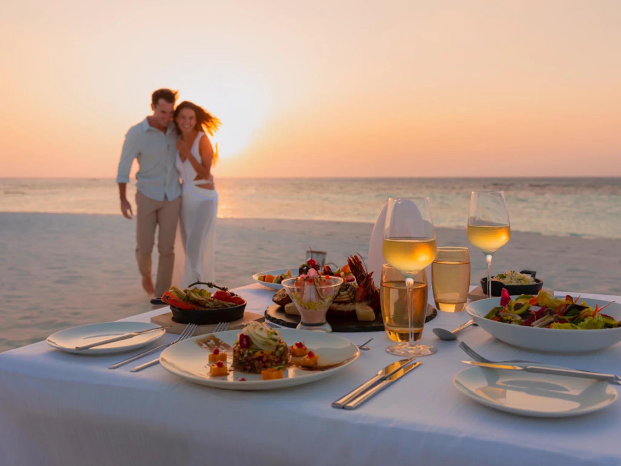 A couple walking along the beach at sunset beside a table set for two, with a seafood platter, salads and glasses of white wine already laid out