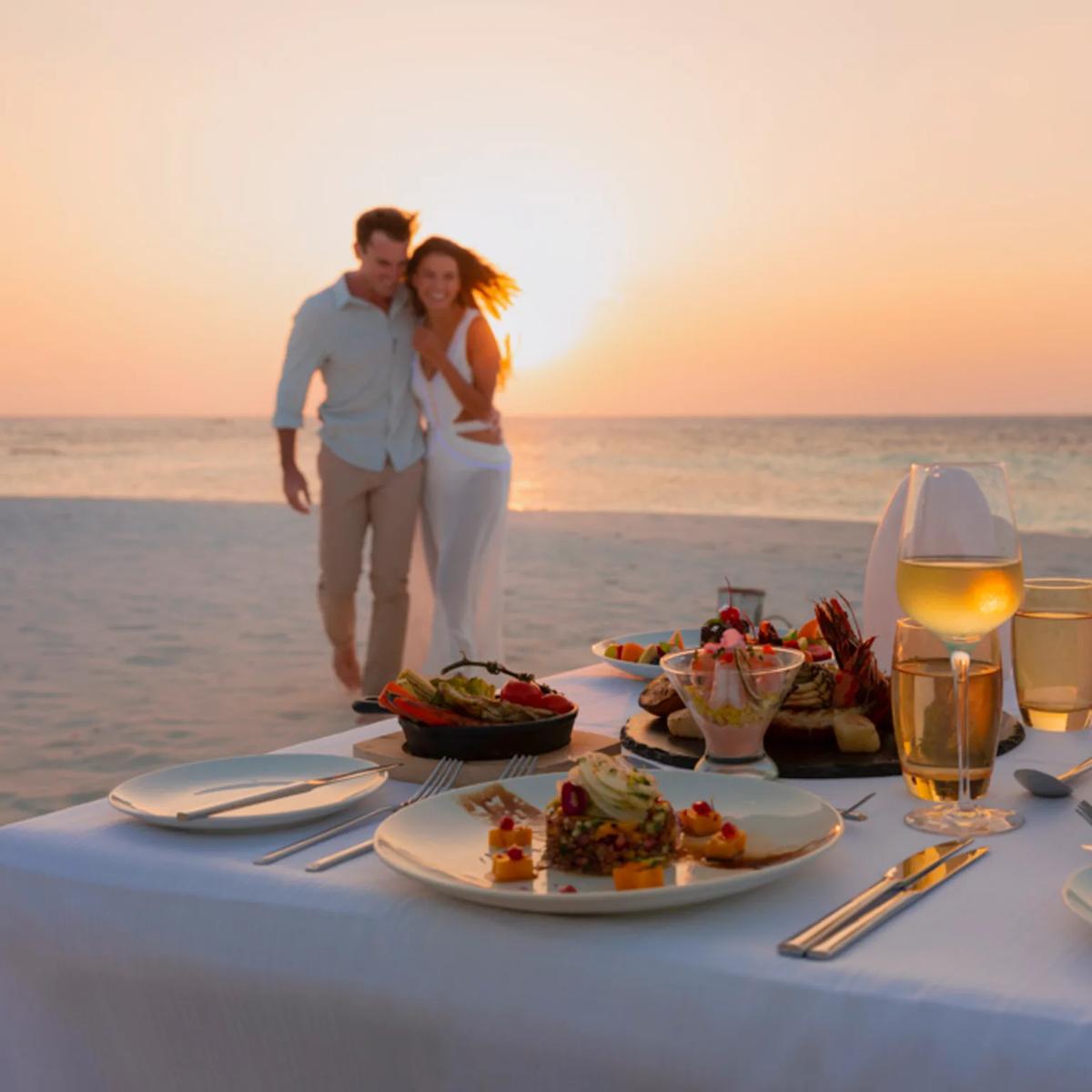 A couple walking along the beach at sunset beside a table set for two, with a seafood platter, salads and glasses of white wine already laid out