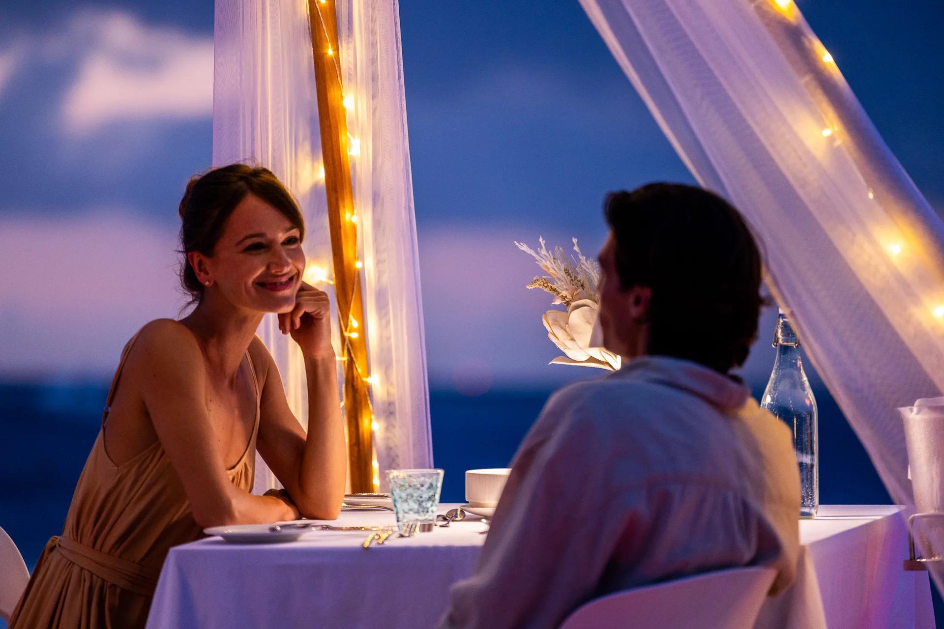 A private candlelit dinner table set up outdoors after dark, sheer curtains and fairy lights framing the scene
