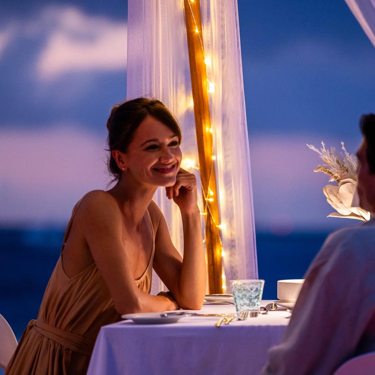 A private candlelit dinner table set up outdoors after dark, sheer curtains and fairy lights framing the scene