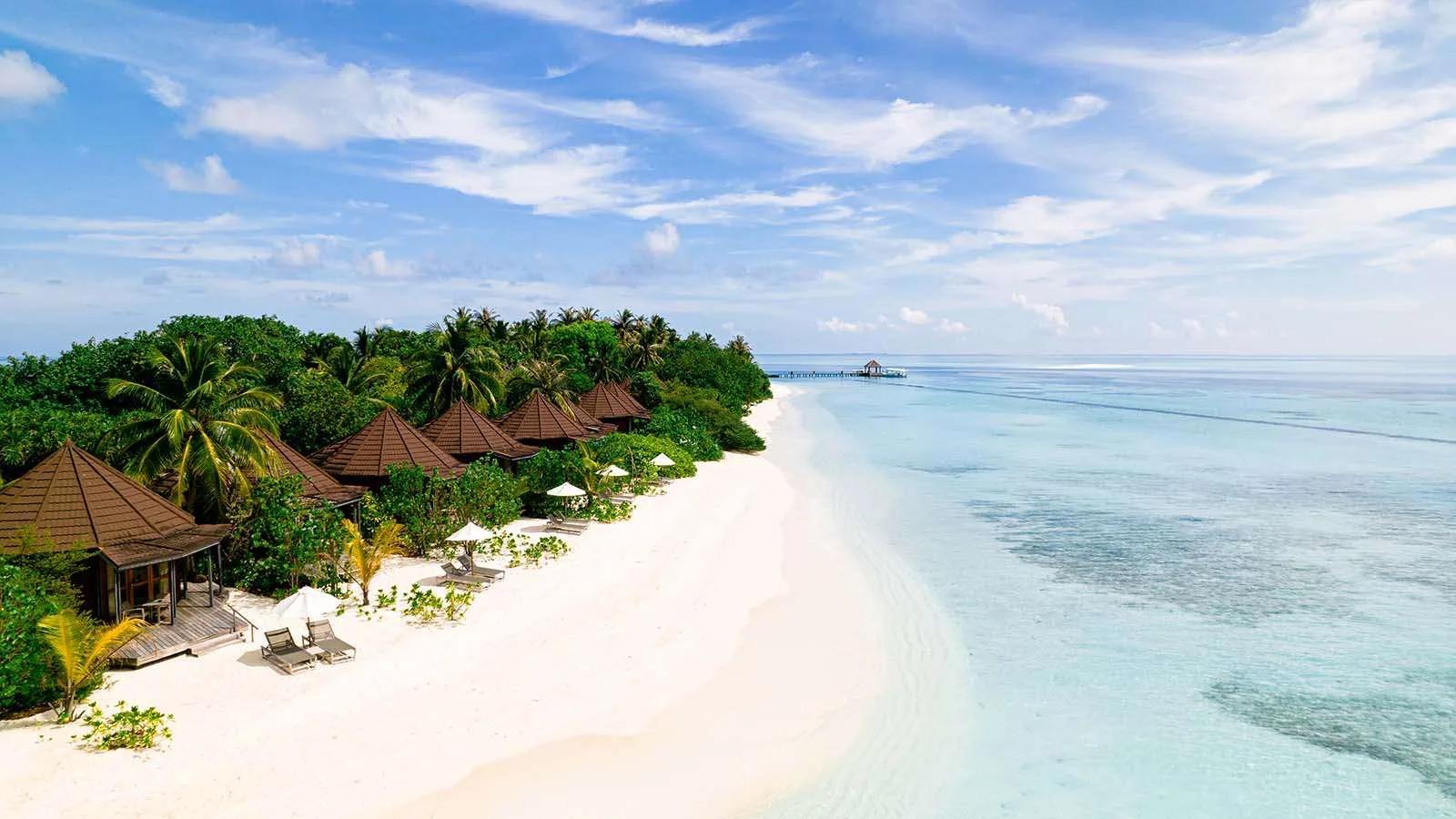 A row of Premium Jacuzzi Beach Villas along the beach, thatched roofs among palm trees with a jetty visible in the distance