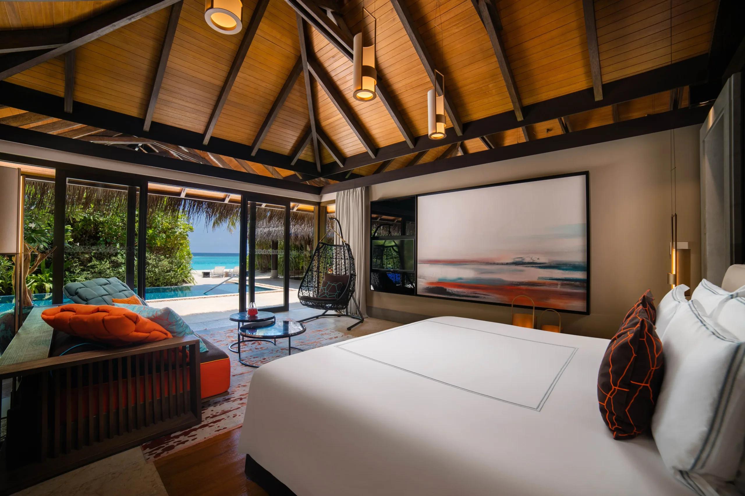 A Beach Pool Villa bedroom with a hanging chair and lounge area, glass doors open to the pool and lagoon beyond