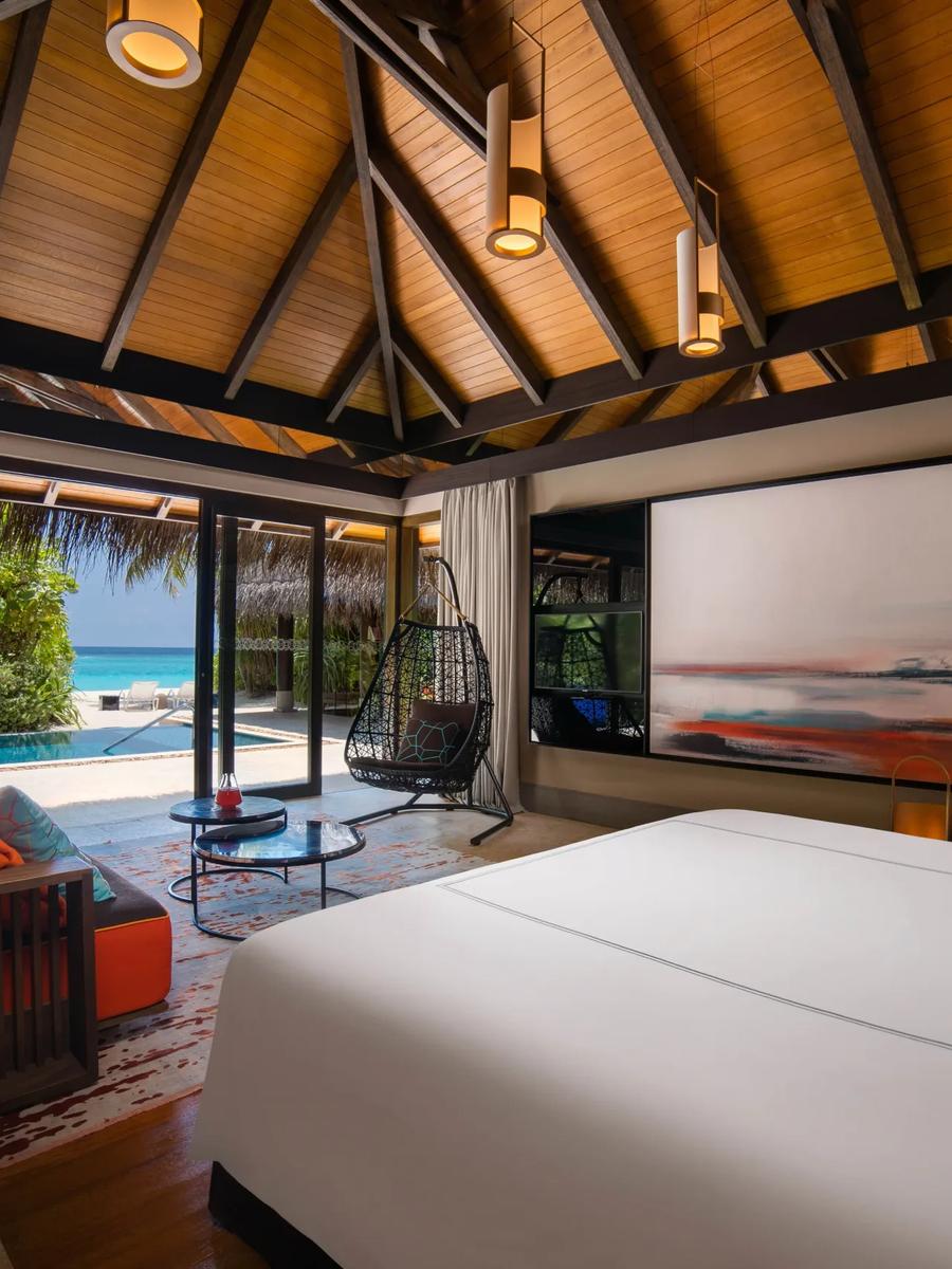 A Beach Pool Villa bedroom with a hanging chair and lounge area, glass doors open to the pool and lagoon beyond
