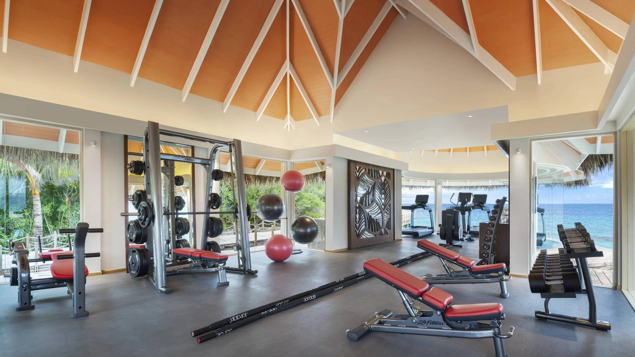 The overwater Fitness Center, weight benches and cable machines facing floor-to-ceiling windows over the lagoon
