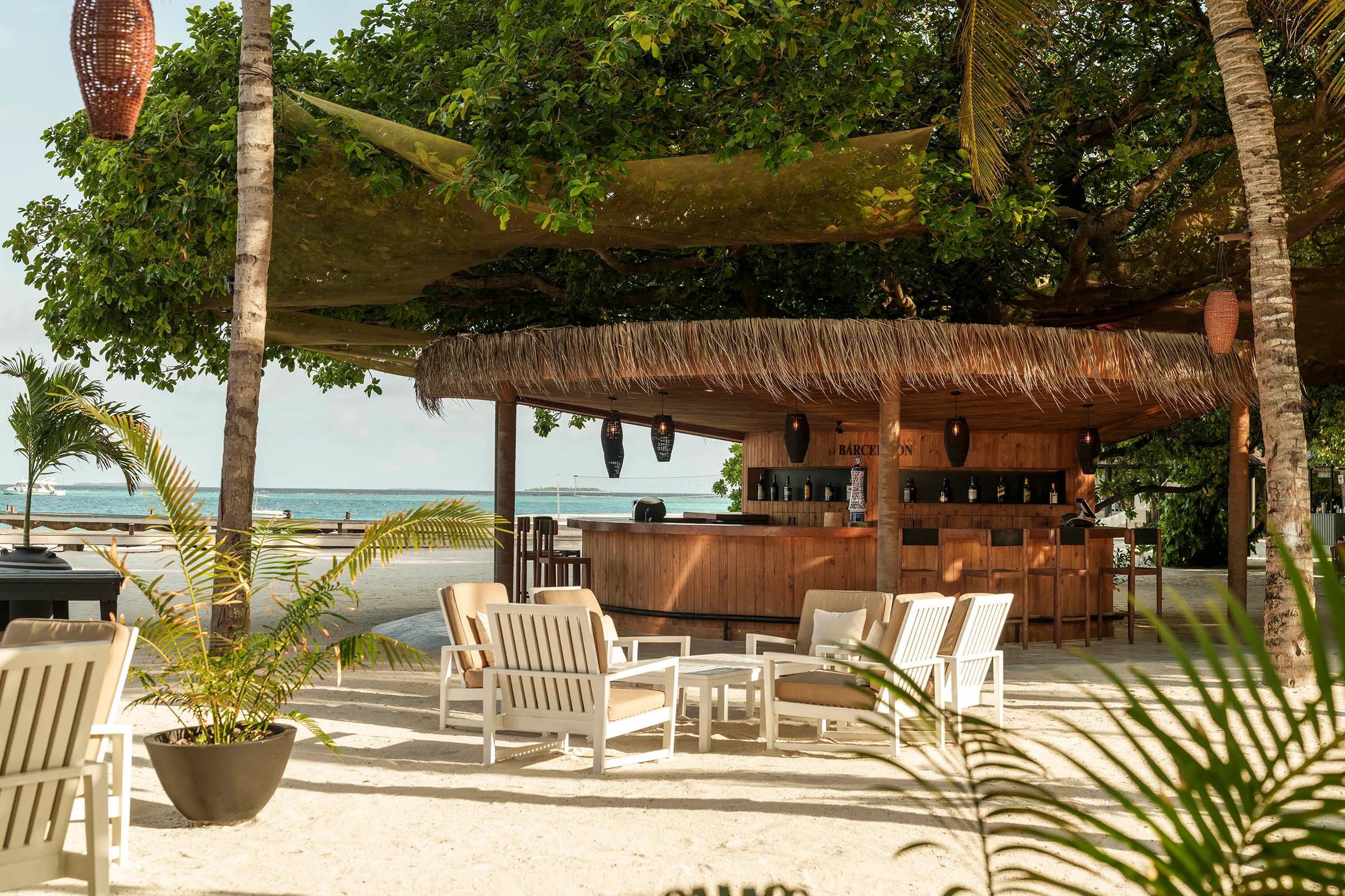 A wooden bar counter stocked with bottles beneath a thatched roof, with lounge seating open to the beach at Waters Edge