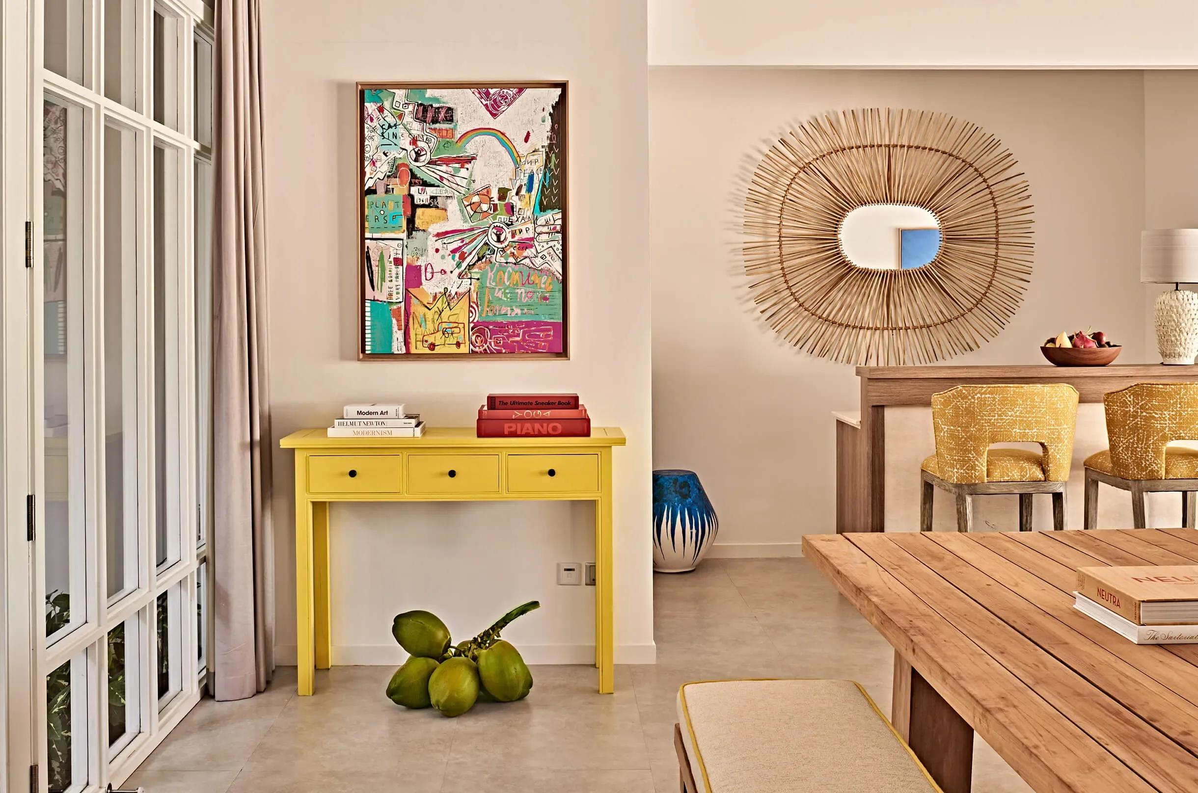 A Two Bedroom Beach Villa's colourful open-plan living space, with a yellow console table under a graffiti-style artwork and a sunburst rattan mirror beyond