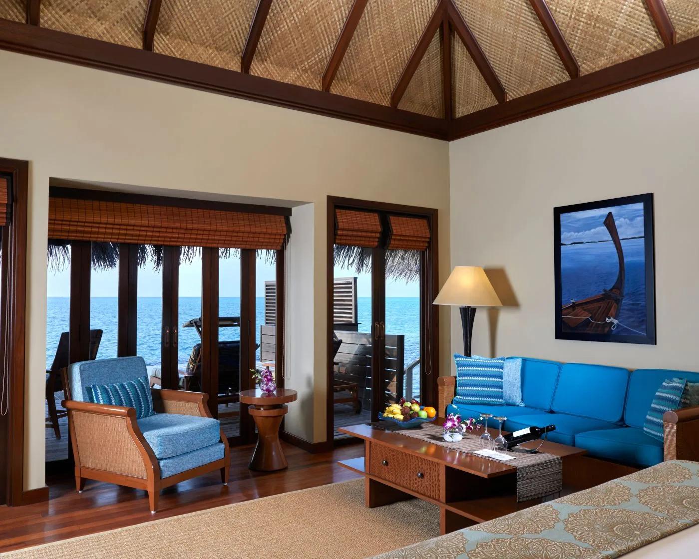 Inside an Overwater Villa with Pool, a blue sectional sofa between two sets of glass doors opening onto the lagoon