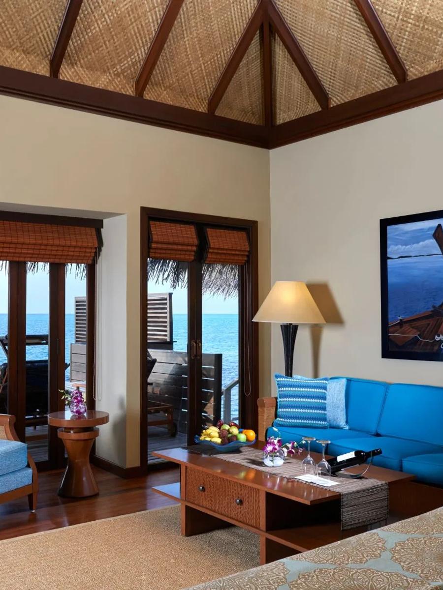 Inside an Overwater Villa with Pool, a blue sectional sofa between two sets of glass doors opening onto the lagoon