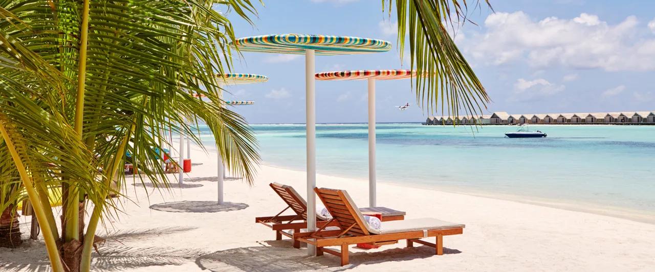 Striped umbrellas and wooden loungers on the beach, with the overwater villa row visible across the lagoon