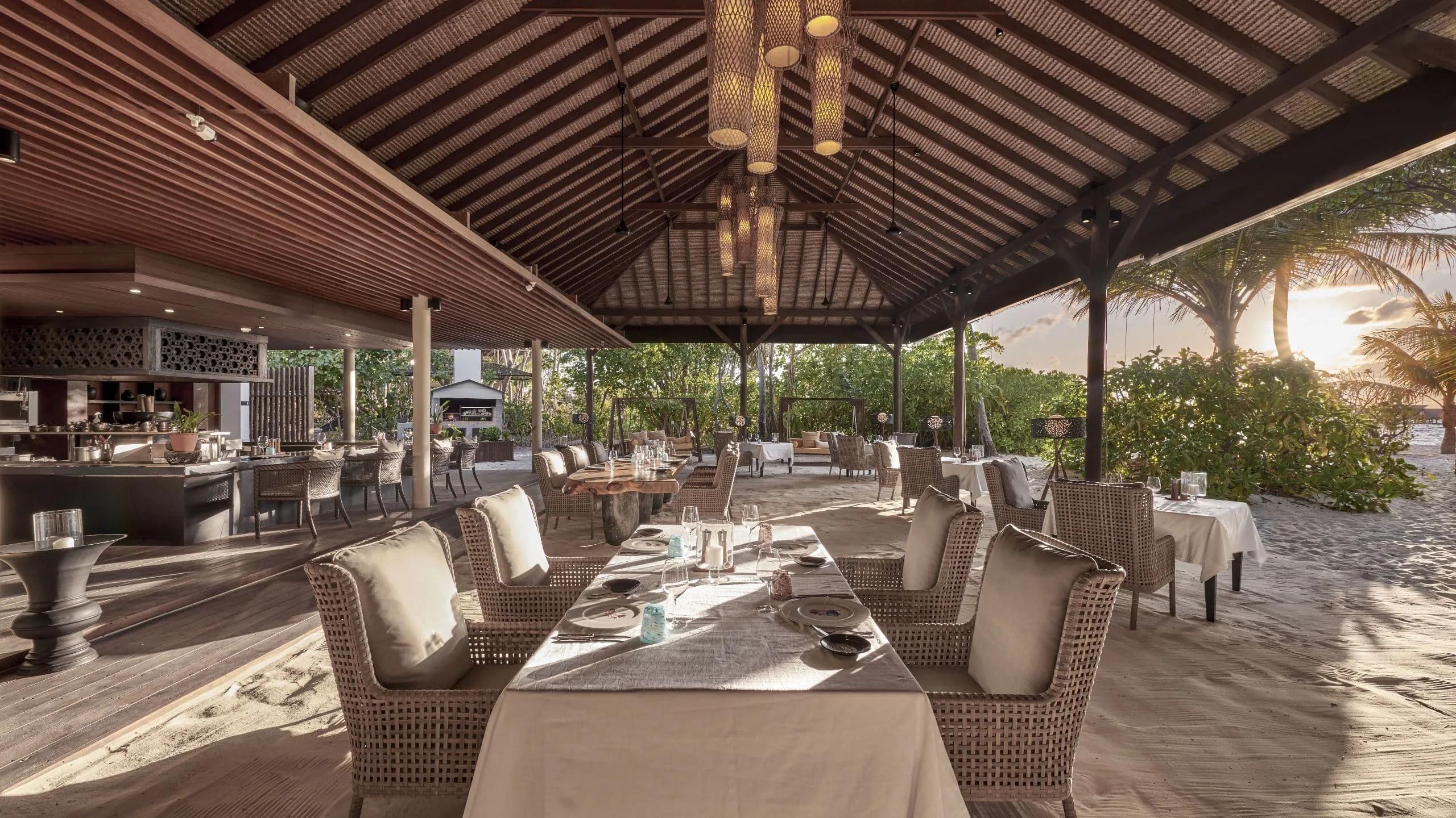 The Island Grill's open-air dining room, tables set in the sand beneath a thatched roof at sunset