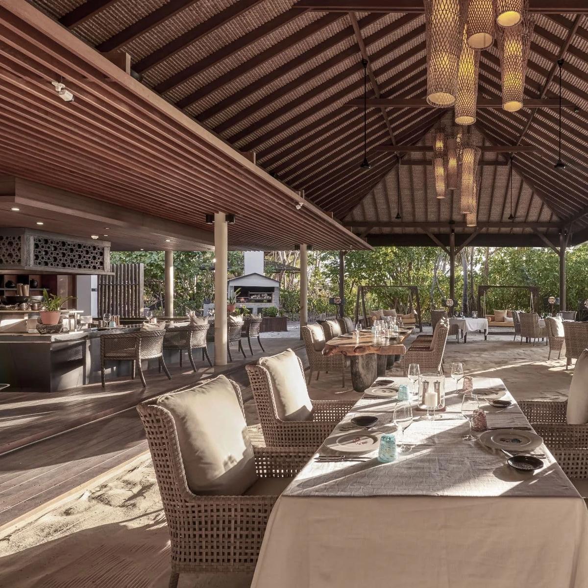 The Island Grill's open-air dining room, tables set in the sand beneath a thatched roof at sunset