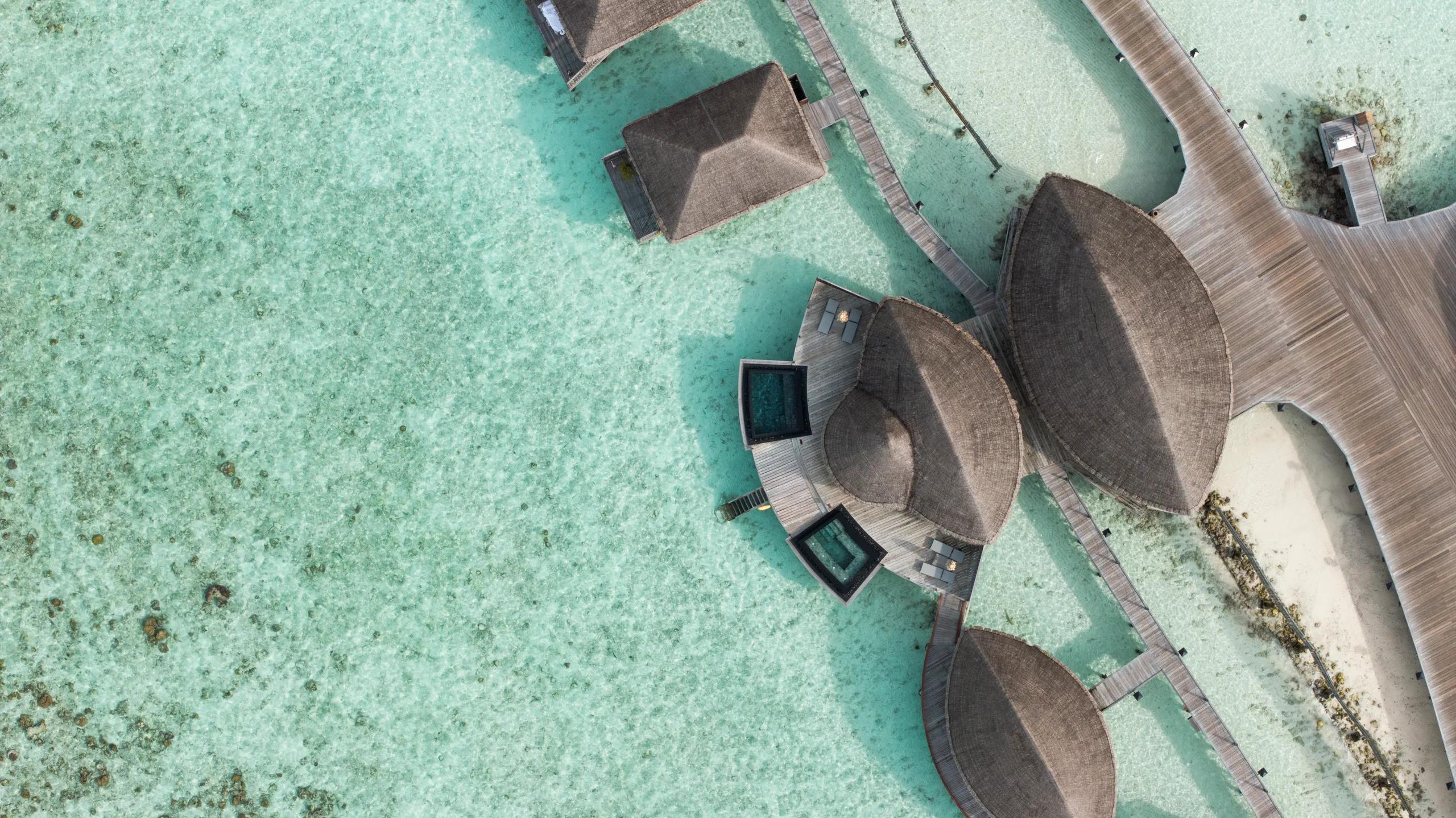 Aerial view of Constance Spa's thatched overwater treatment pavilions on stilts above the lagoon, connected by wooden jetties, with plunge pools visible on the decks