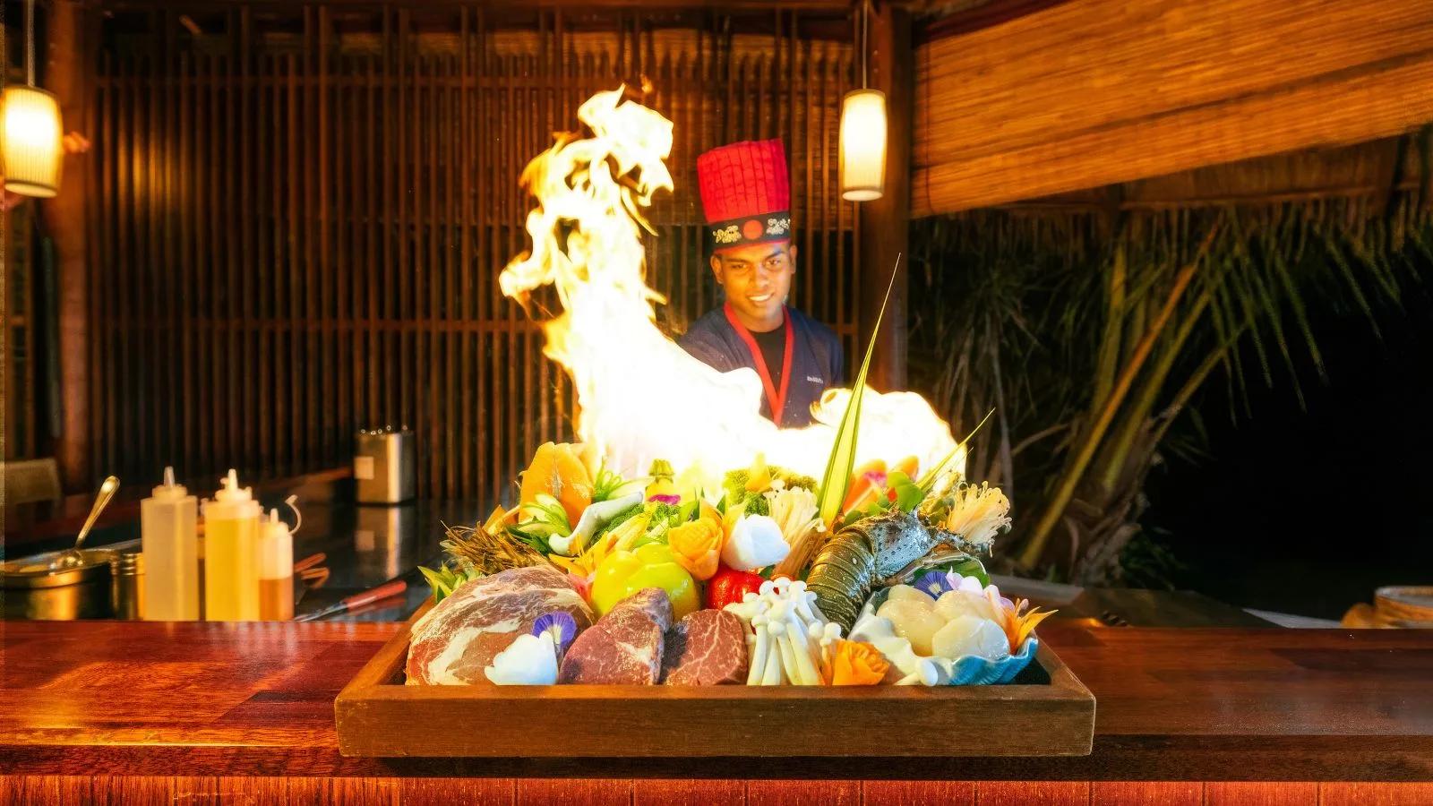 A chef sends flames up from a teppanyaki grill loaded with lobster, beef, and vegetables