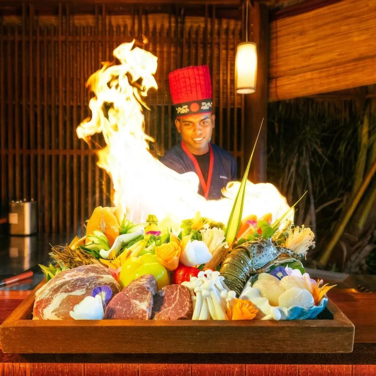 A chef sends flames up from a teppanyaki grill loaded with lobster, beef, and vegetables