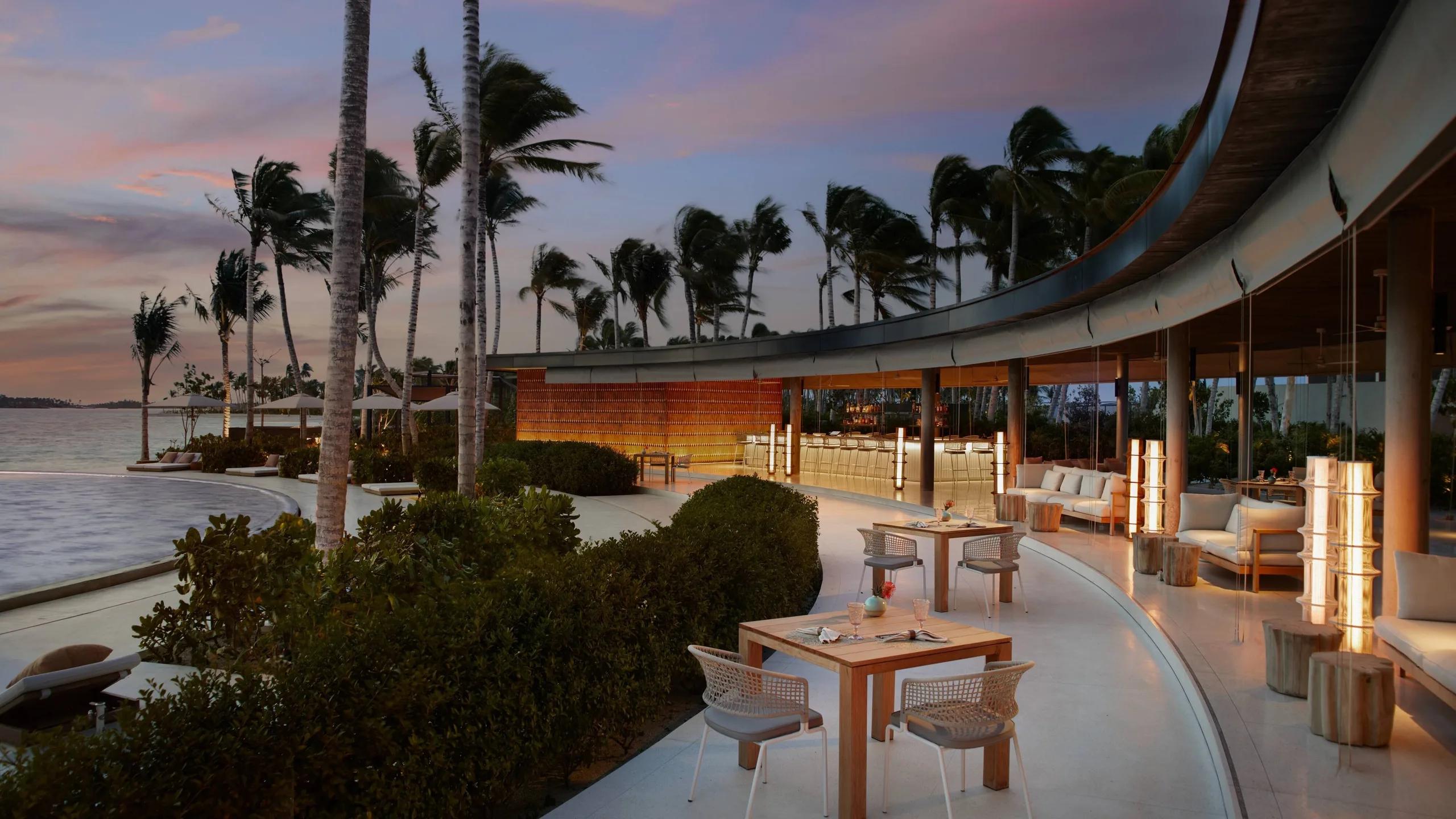 EAU Bar's curved terrace lit up at dusk, palm trees silhouetted against a pink sky