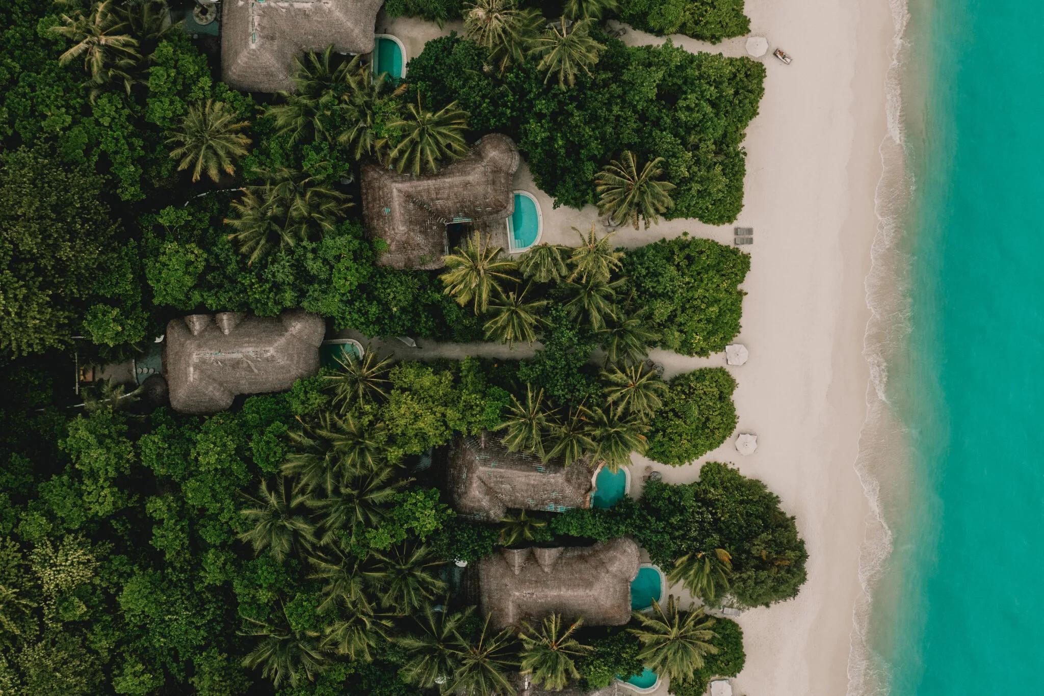 An aerial view along Kunfunadhoo's beach, showing several villas' thatched roofs and private pools among the jungle, with the reef beyond — representative of the island's beachfront villa categories; no dedicated photograph of the nine-bedroom Villa 11 Residence was available in this research batch