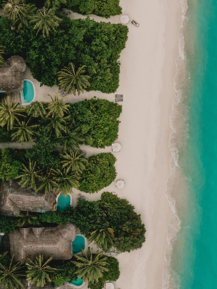 An aerial view along Kunfunadhoo's beach, showing several villas' thatched roofs and private pools among the jungle, with the reef beyond — representative of the island's beachfront villa categories; no dedicated photograph of the nine-bedroom Villa 11 Residence was available in this research batch