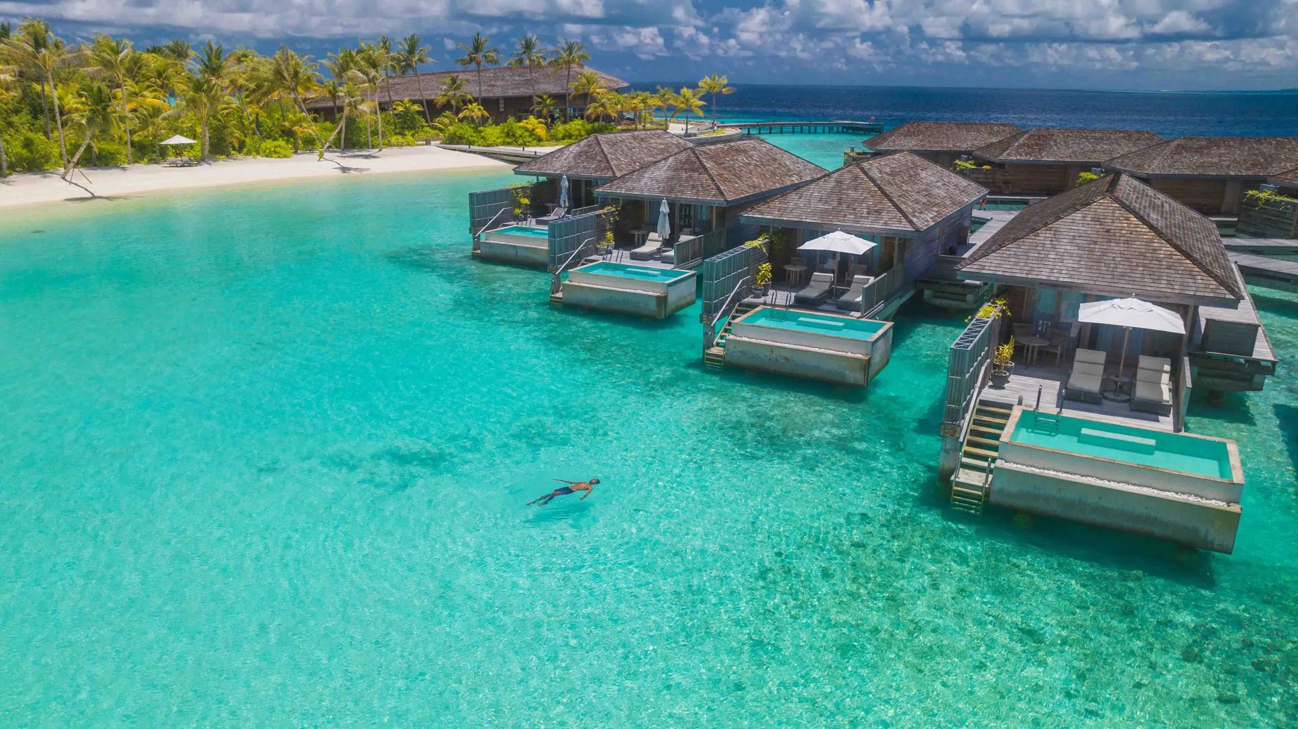 A wide aerial view of Kagi Maldives — white sand beach, dense palm cover, and a row of overwater villas reaching out to a jetty over the turquoise lagoon