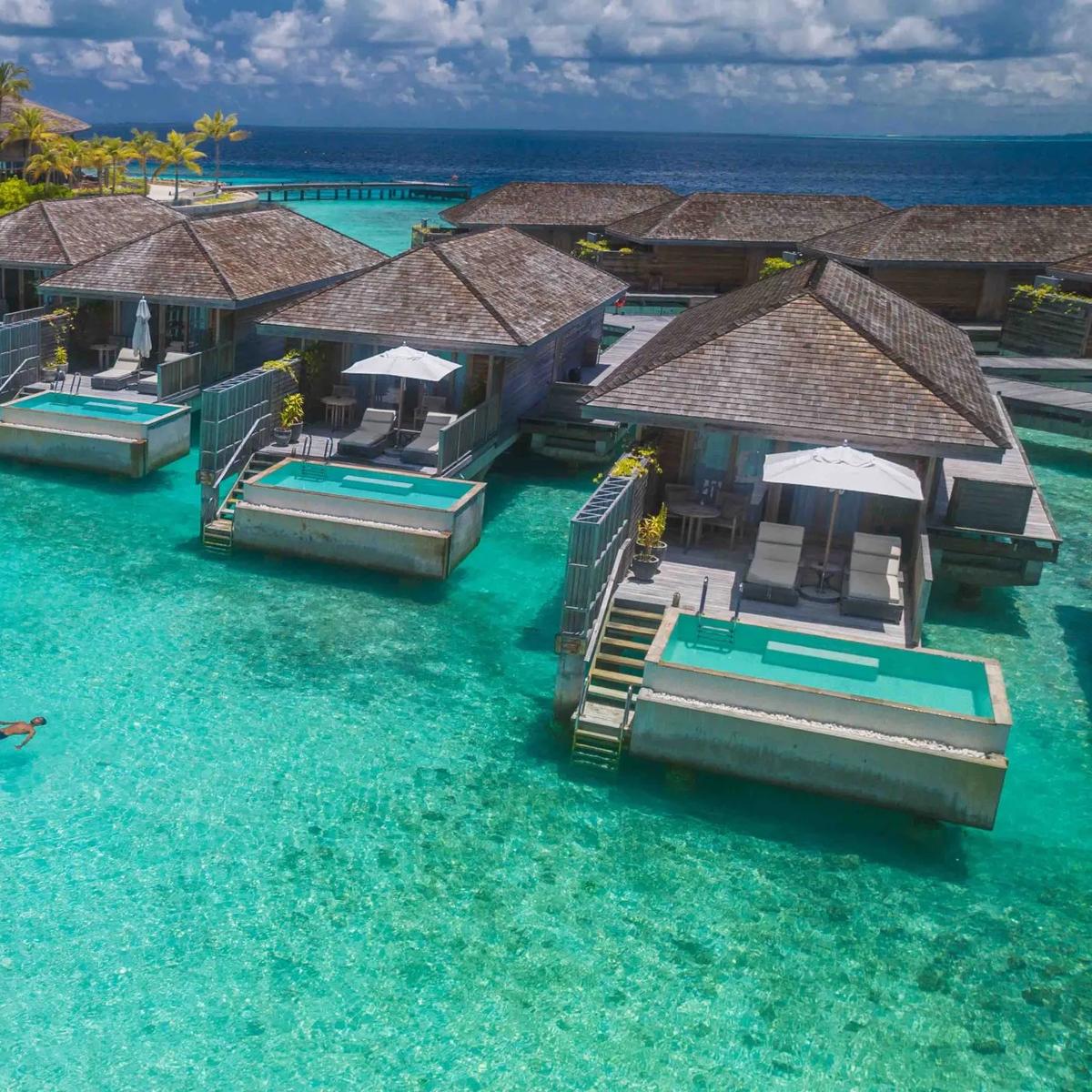 A wide aerial view of Kagi Maldives — white sand beach, dense palm cover, and a row of overwater villas reaching out to a jetty over the turquoise lagoon