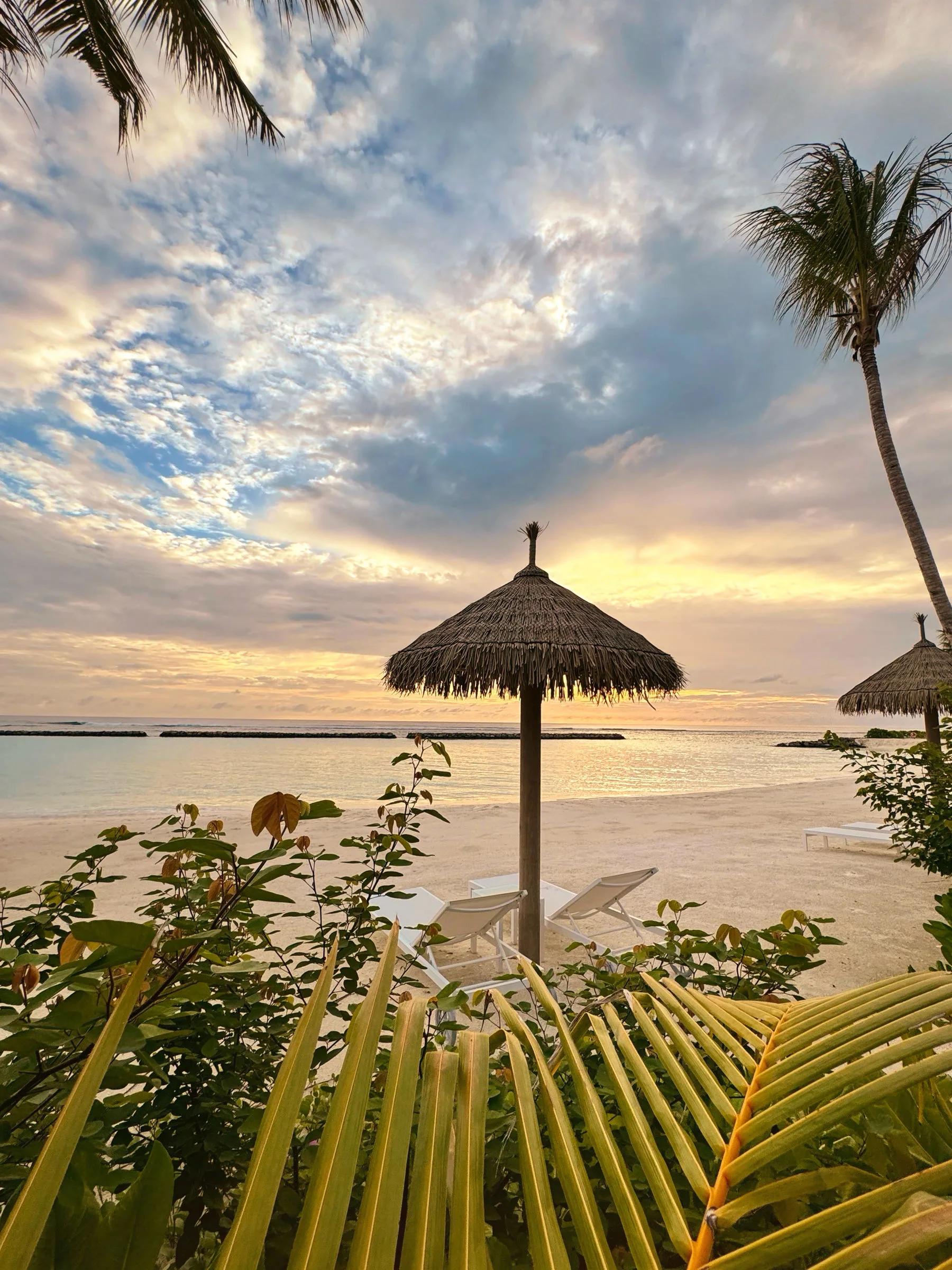 A thatched beach umbrella and two loungers on white sand at sunset, palm trees framing the scene