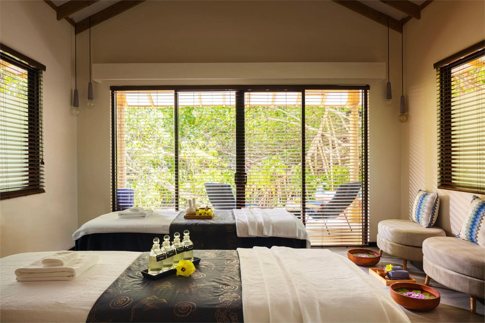 Two massage beds laid with dark patterned linens and treatment oils in the resort's spa, floor-to-ceiling windows opening onto greenery and a daybed beyond