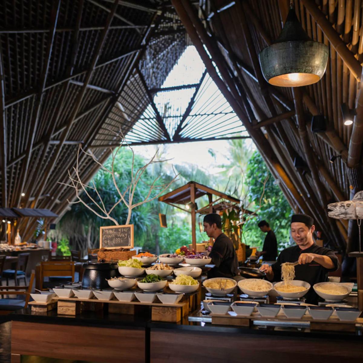 Chefs preparing noodles at ONU Marché's live cooking station, bowls of fresh vegetables and garnishes laid out under the restaurant's bamboo roof
