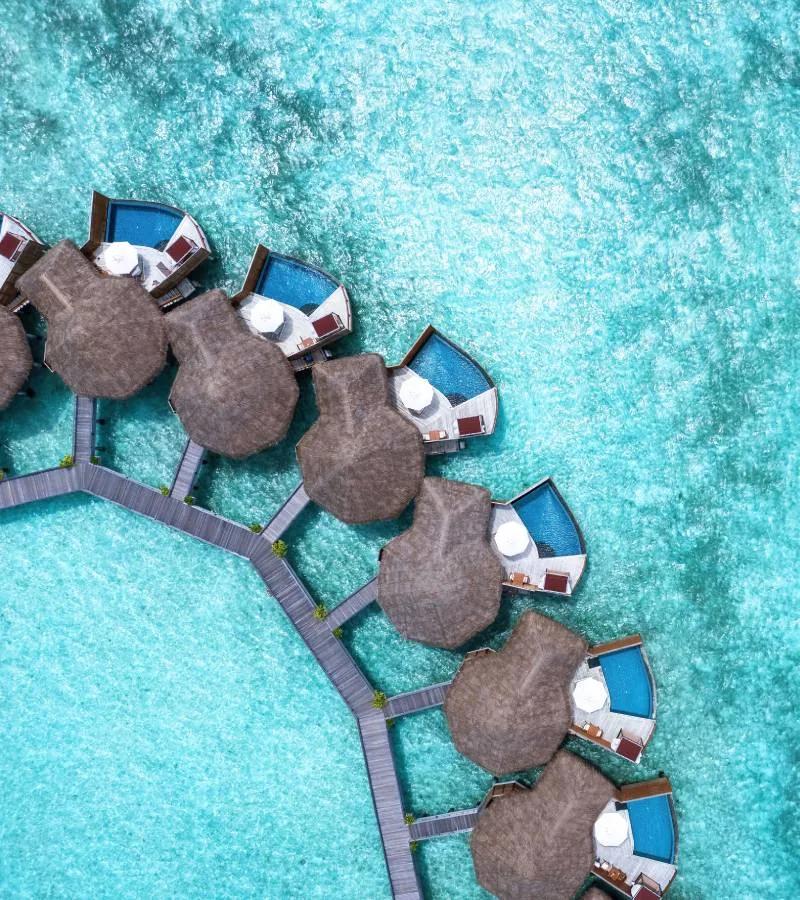 Directly overhead view of Baros's thatched-roof Water Pool Villas with private plunge pools, connected by a timber jetty over turquoise water