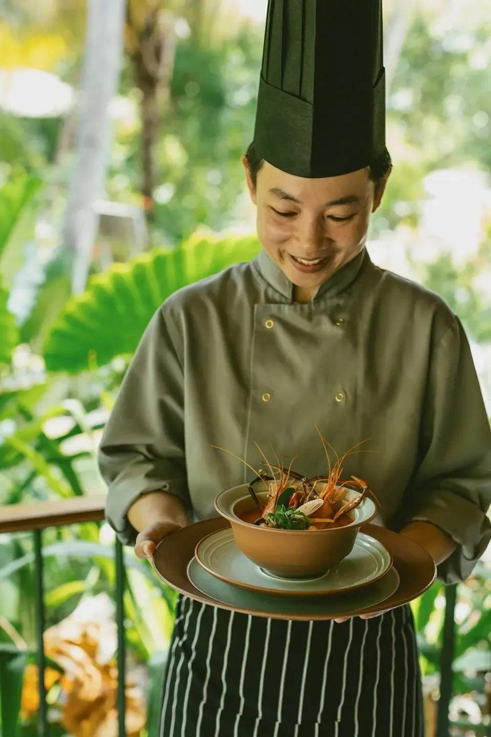 A chef presenting a bowl of Thai tom yum-style soup with prawns, garnished tableside
