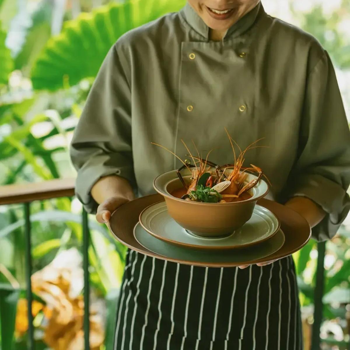 A chef presenting a bowl of Thai tom yum-style soup with prawns, garnished tableside