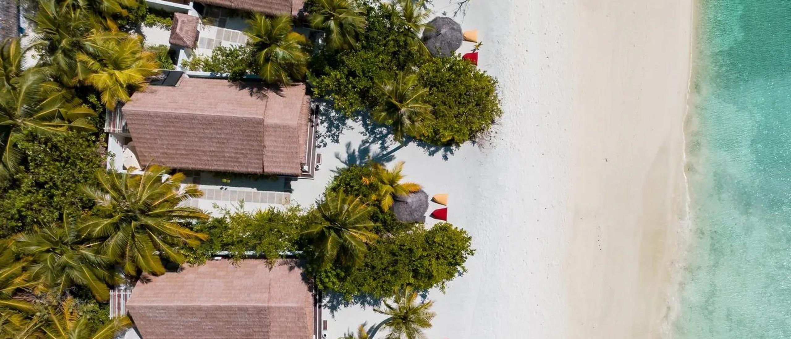 Aerial view of beachfront villas along Maadhoo Island's white-sand beach, thatched umbrellas spaced along the tree line