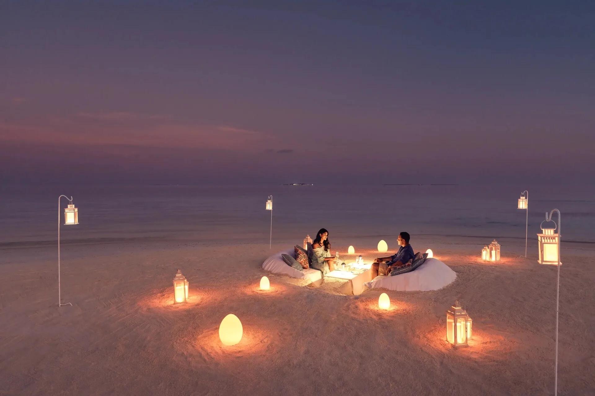 A couple seated at a private dinner table on the beach at dusk, ringed by lit lanterns