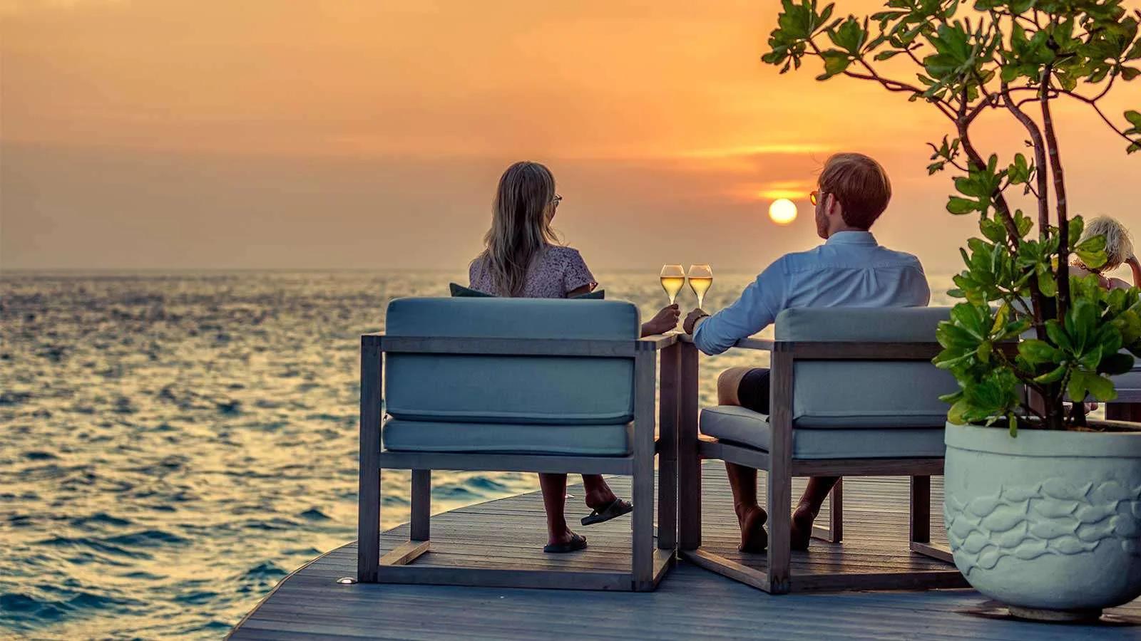 A couple toasting with champagne at the overwater Champagne Pavilion as the sun sets over the ocean