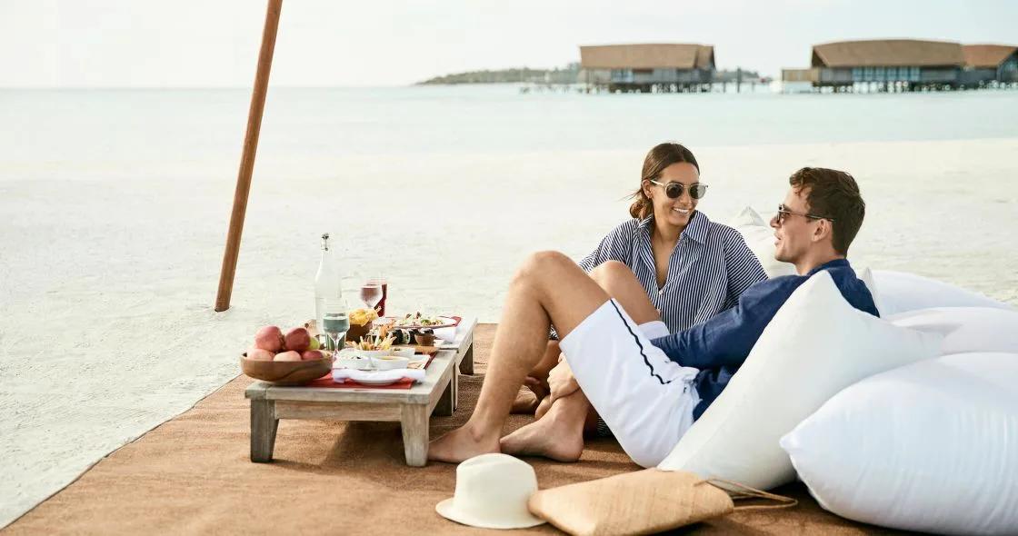A couple sharing a beach picnic on the sand, the resort's overwater villas in view behind them