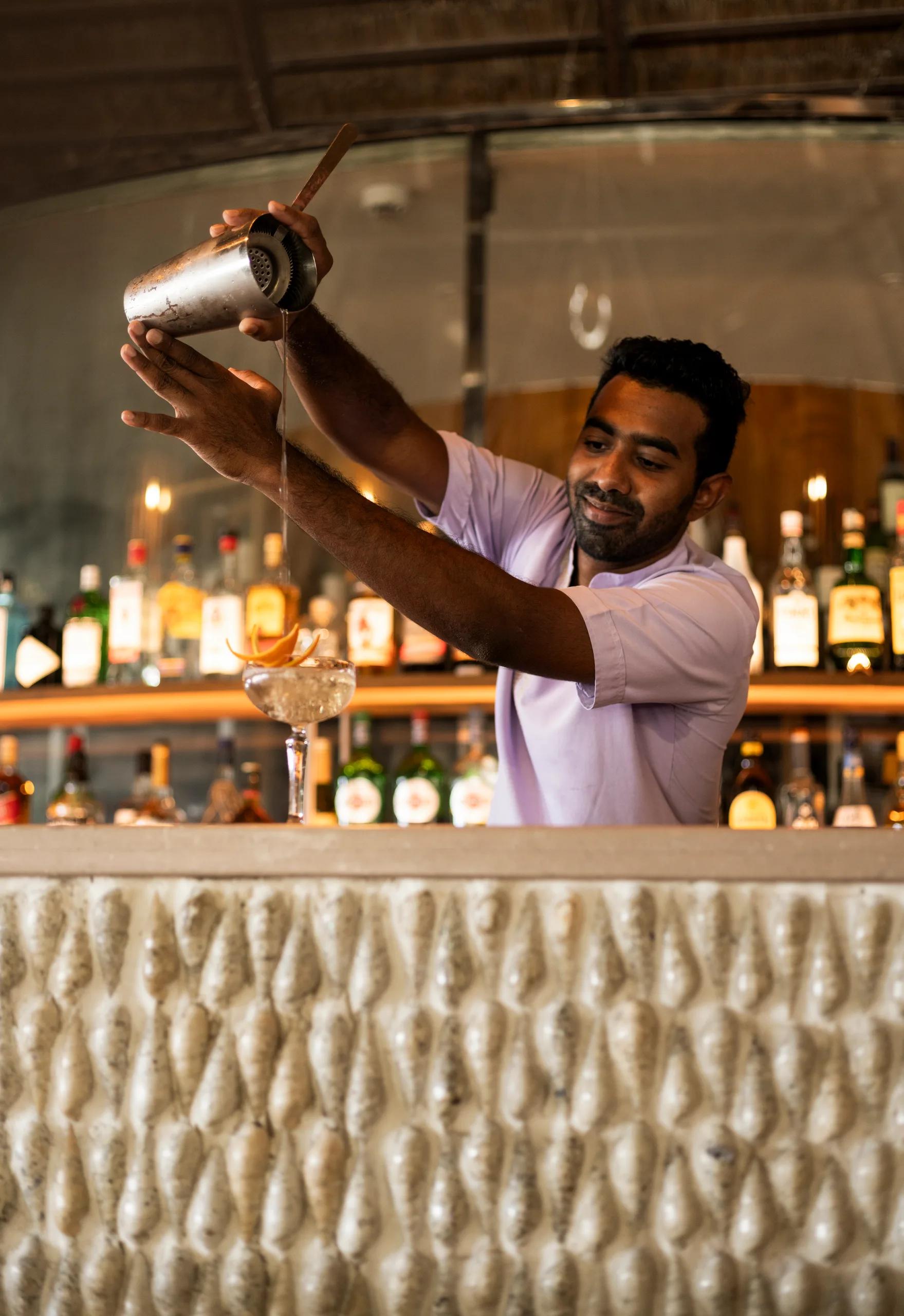 A bartender pouring a shaken cocktail into a coupe glass garnished with an orange twist, bottles lit behind him