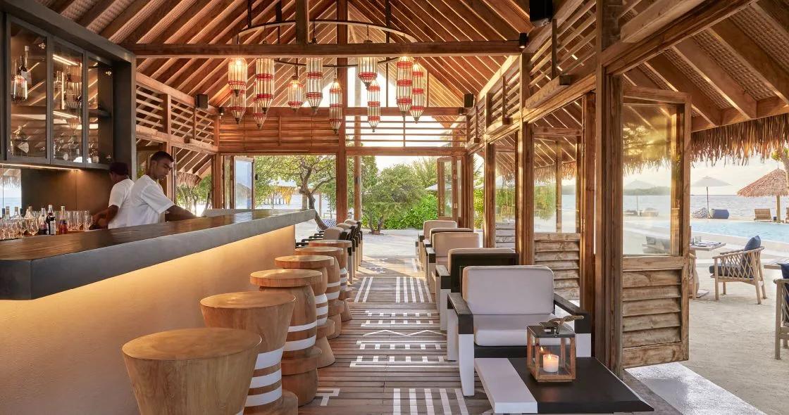 Faru Bar's counter, with a bartender at work and the pool and lagoon visible beyond