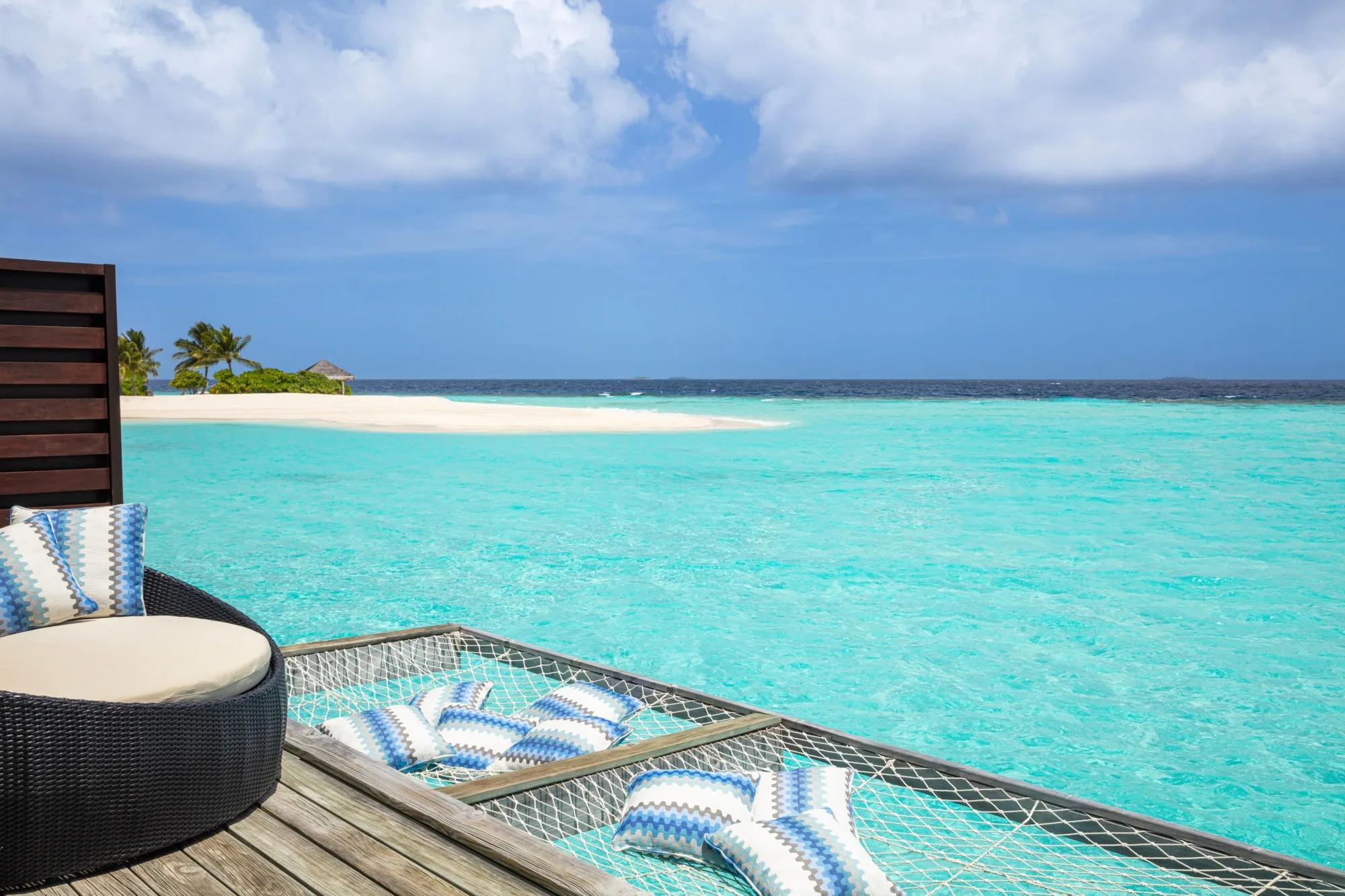 An overwater hammock strung with striped cushions on a private deck above turquoise water, a sandbank and distant island visible ahead