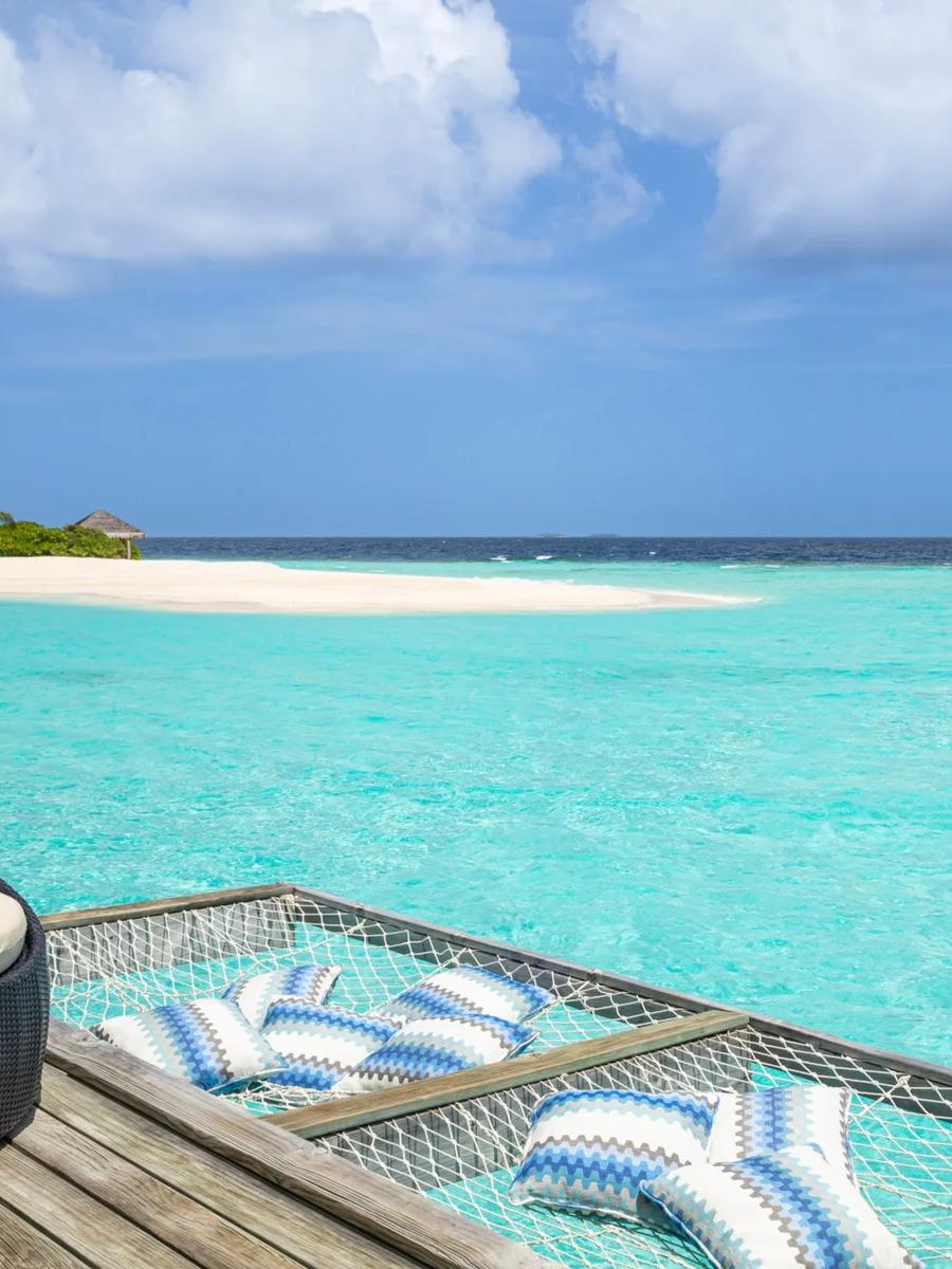 An overwater hammock strung with striped cushions on a private deck above turquoise water, a sandbank and distant island visible ahead