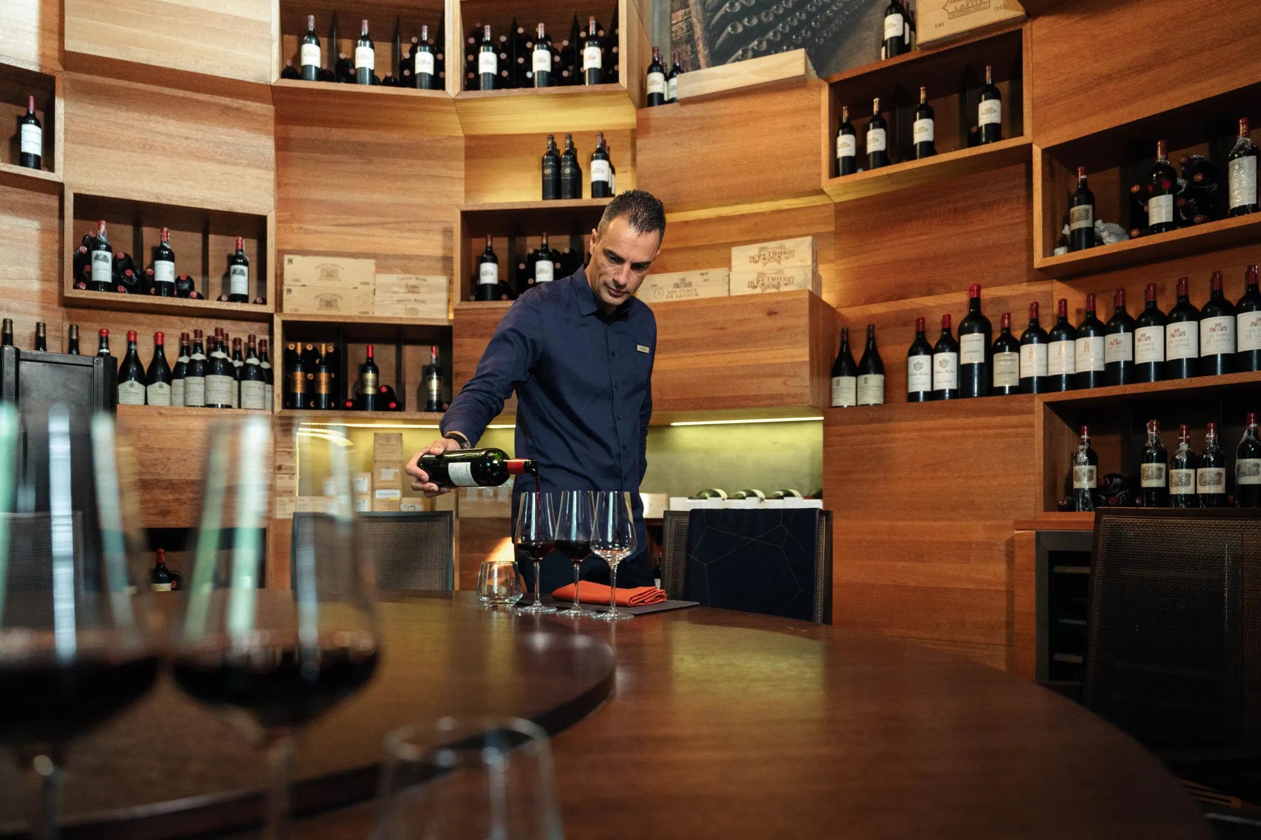 A sommelier pours wine inside Tavaru's cellar, bottles shelved floor to ceiling around the room