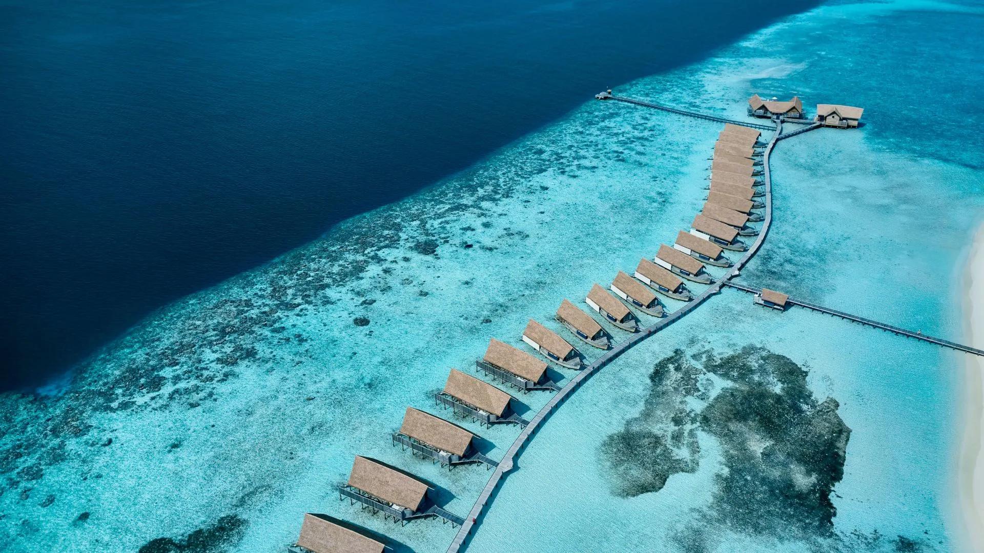 Aerial view of COMO Cocoa Island's overwater villas curving along the reef edge, from deep blue open water into the pale turquoise lagoon