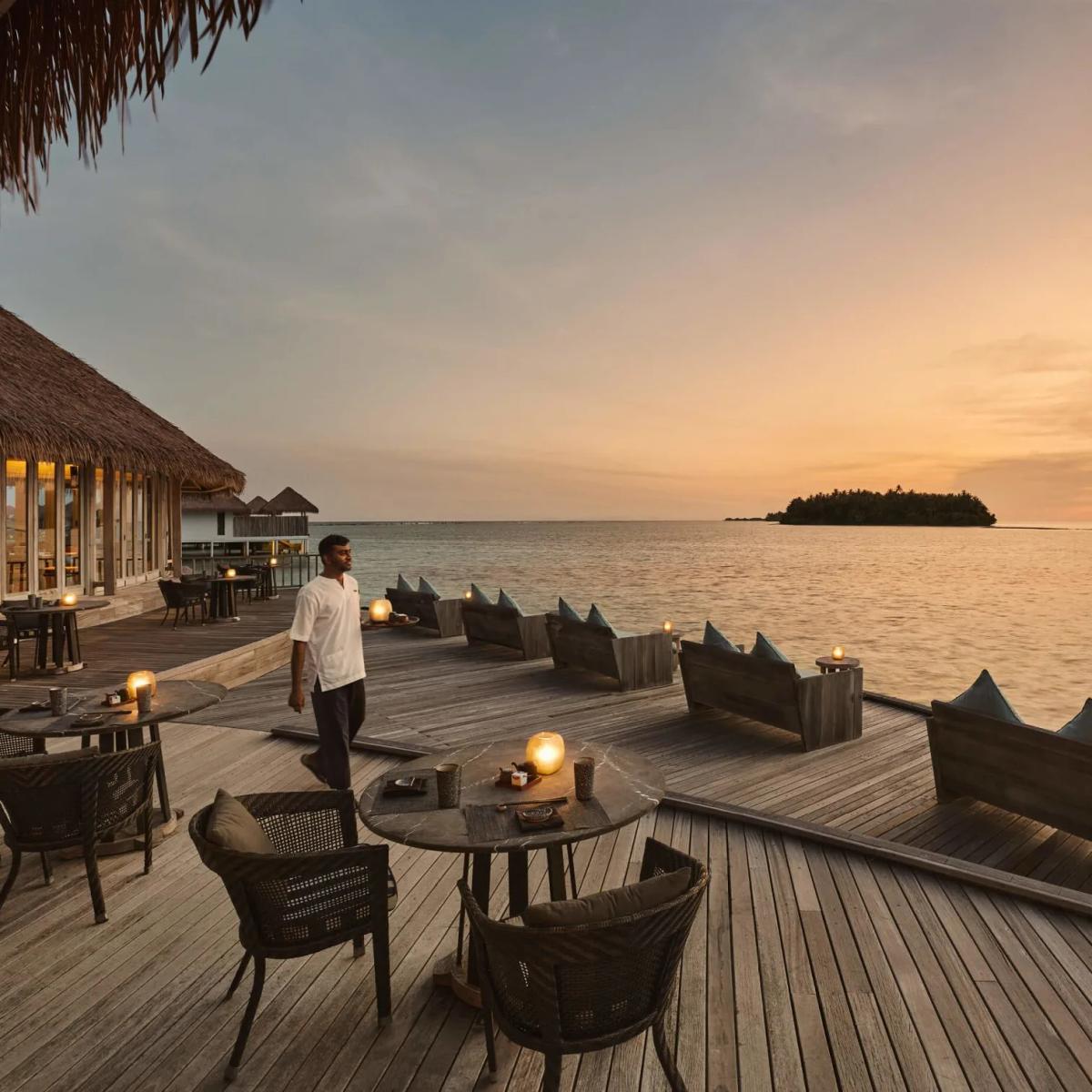 Tai's overwater deck set for dinner at sunset, with a small island silhouetted on the horizon