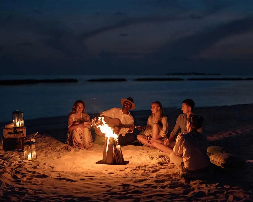 Guests gathered around a beach campfire after dark, one playing guitar.
