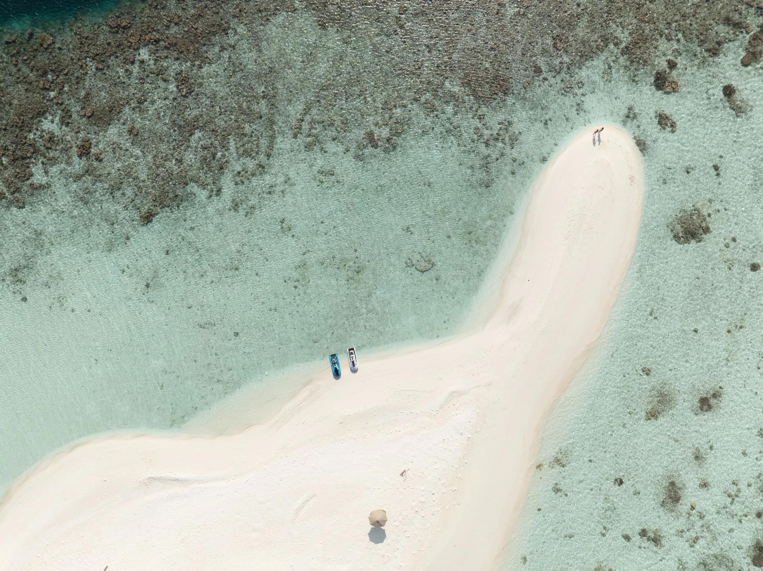 Aerial view of a white sandbank curving into the lagoon at Sun Siyam Olhuveli, reef patterns visible through clear water with two jet cars beached on the sand