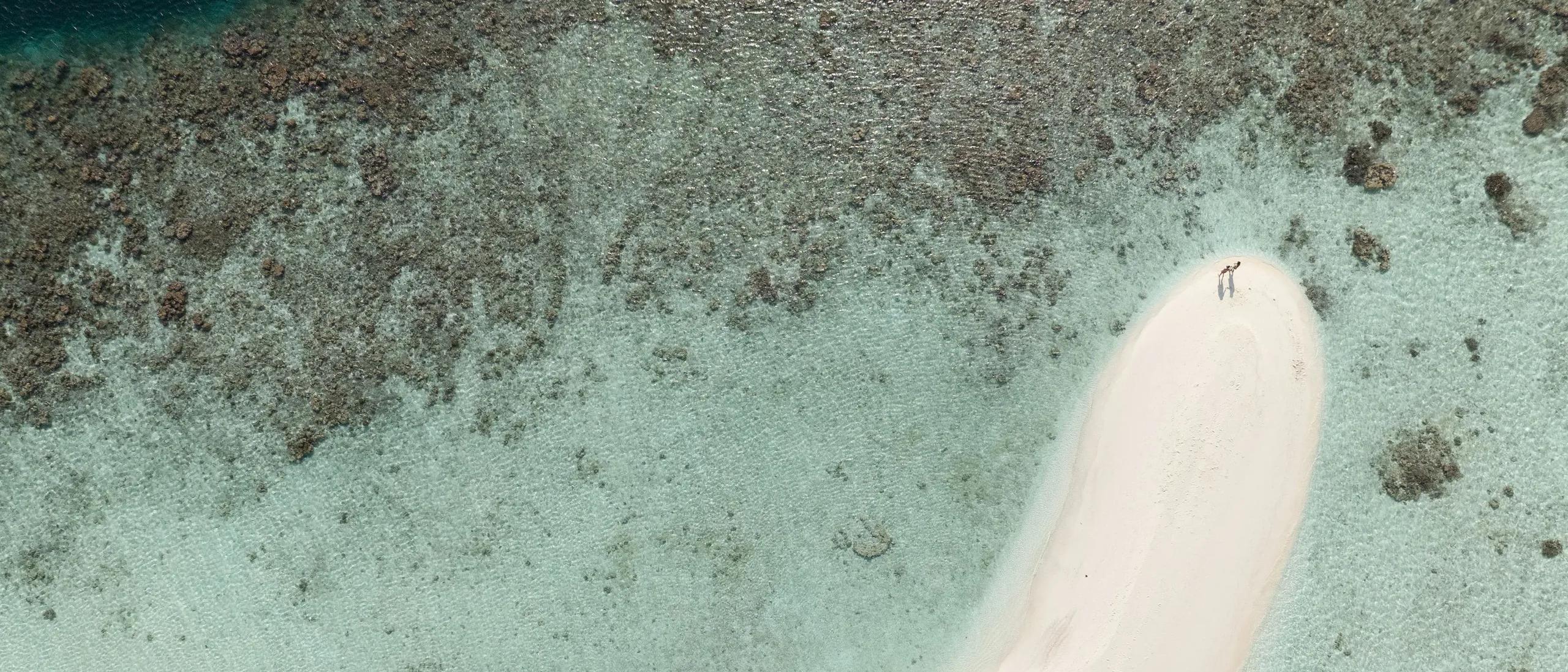 Aerial view of a white sandbank curving into the lagoon at Sun Siyam Olhuveli, reef patterns visible through clear water with two jet cars beached on the sand