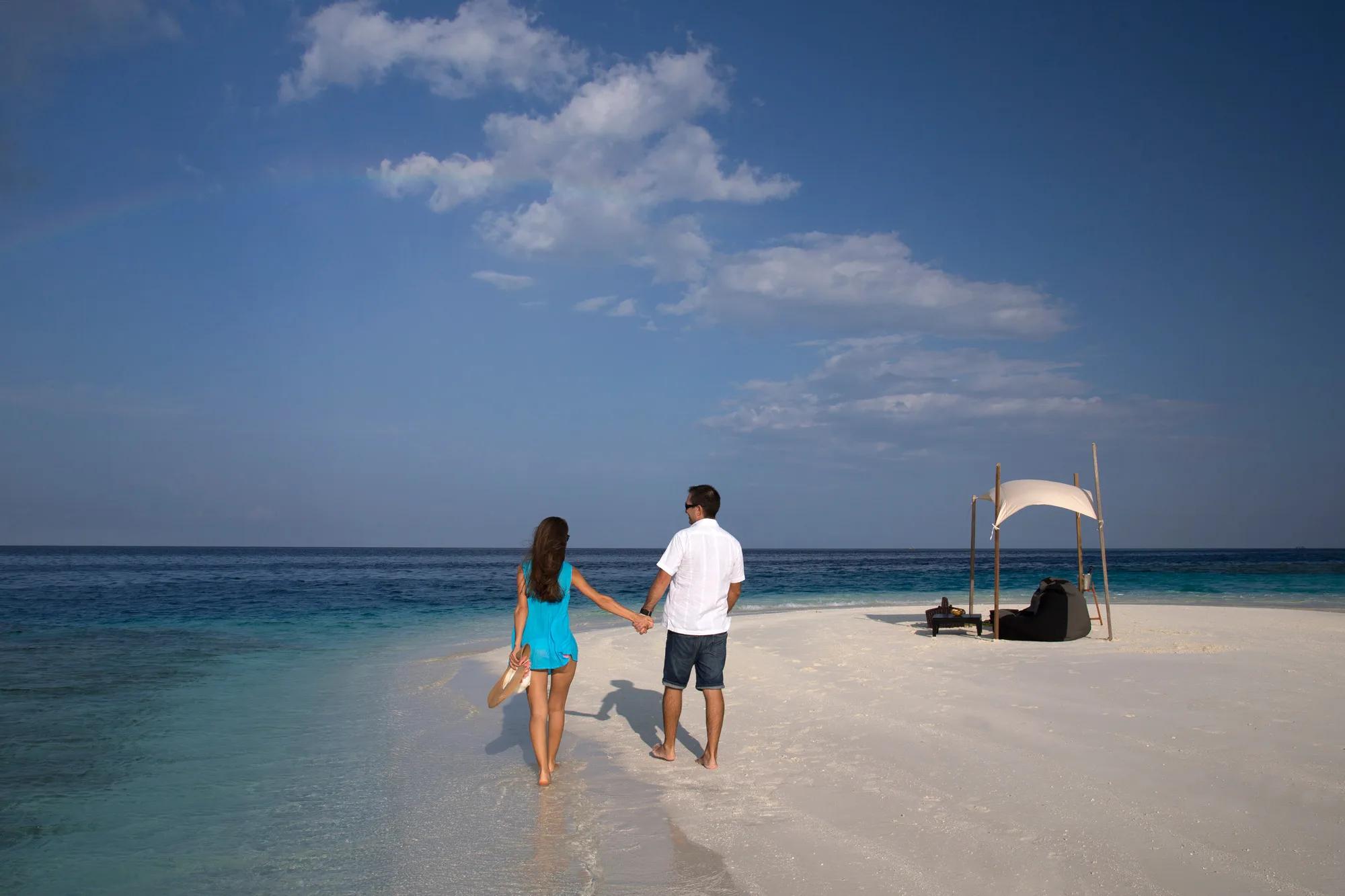 A couple walking hand in hand along a sandbank toward a shaded private picnic setup